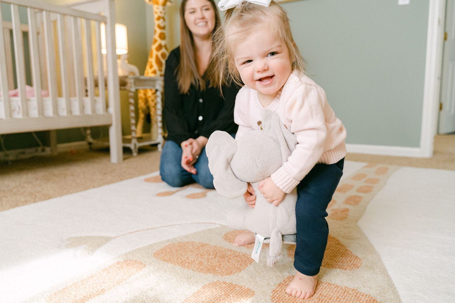 A baby crawls on a carpeted floor while a woman in dark clothing watches from nearby.