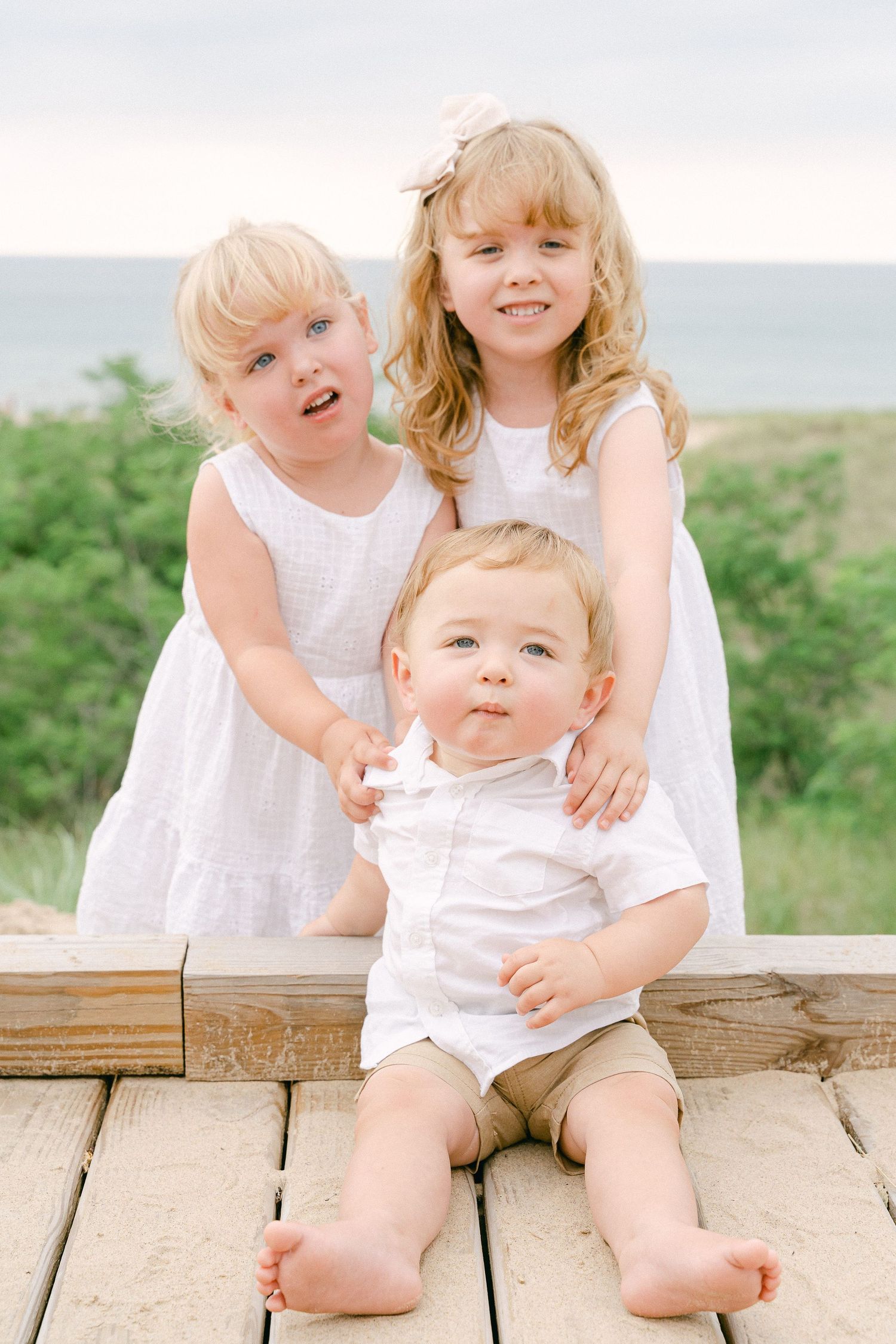 Children in white summer outfits sit together on wooden deck outdoors.