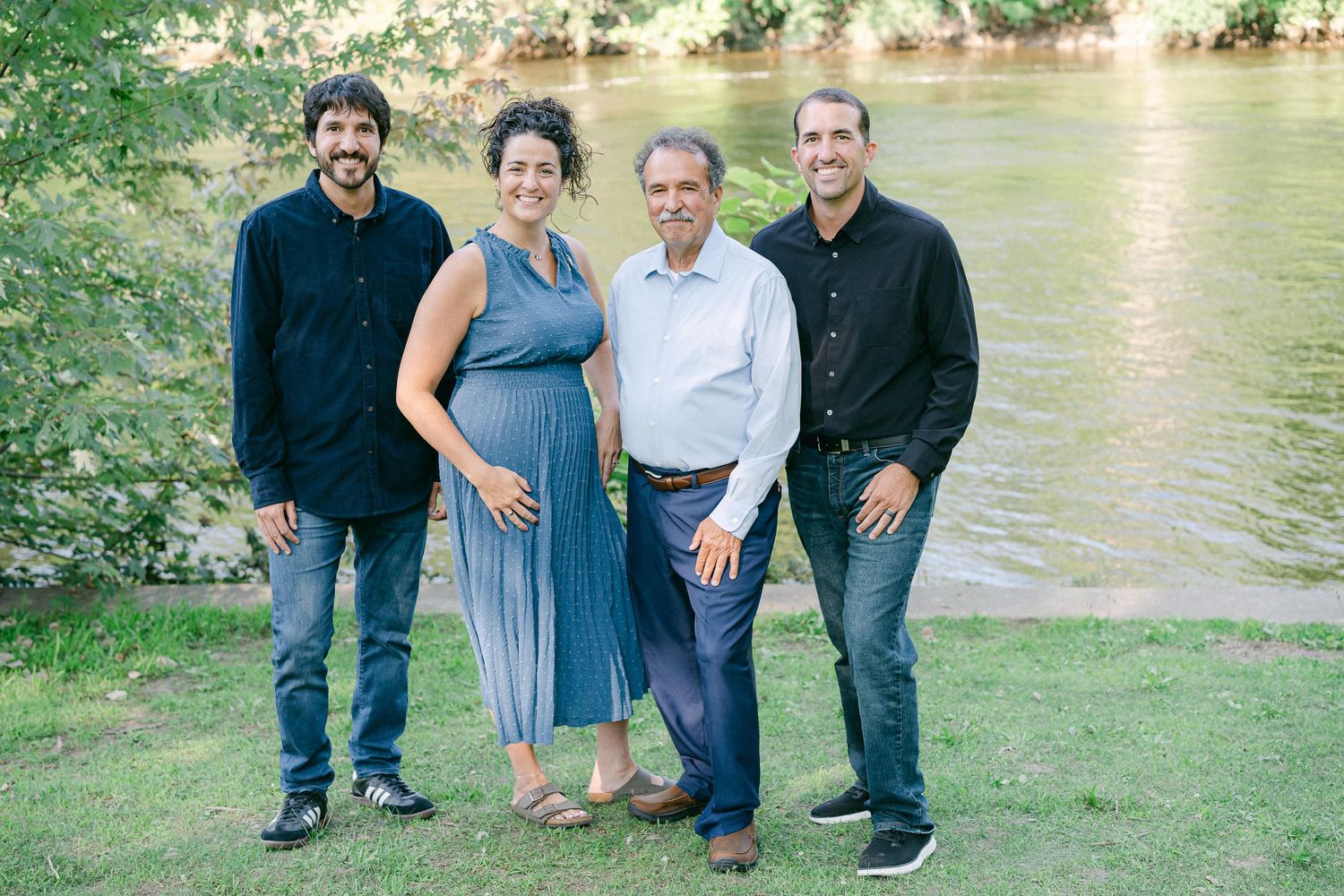 Group of people standing together outdoors near a river bank in a casual outdoor setting.