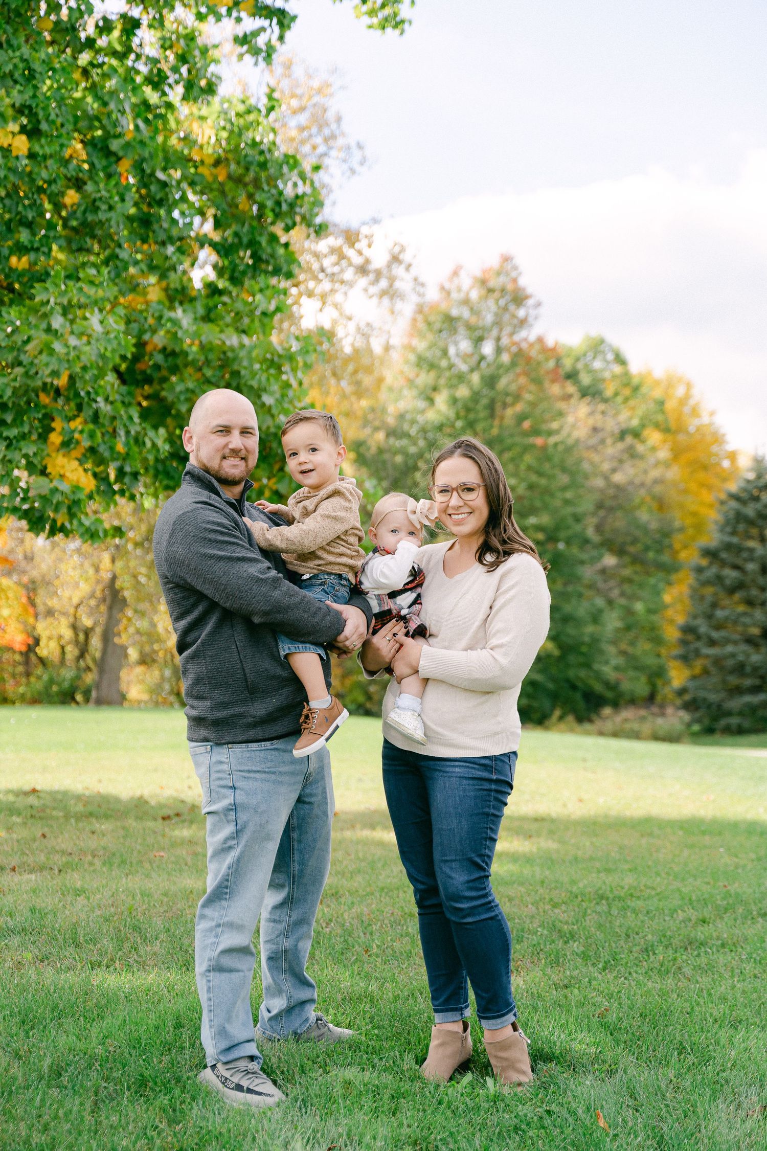 A family stands together on green grass beneath a tree with golden autumn leaves in the background.