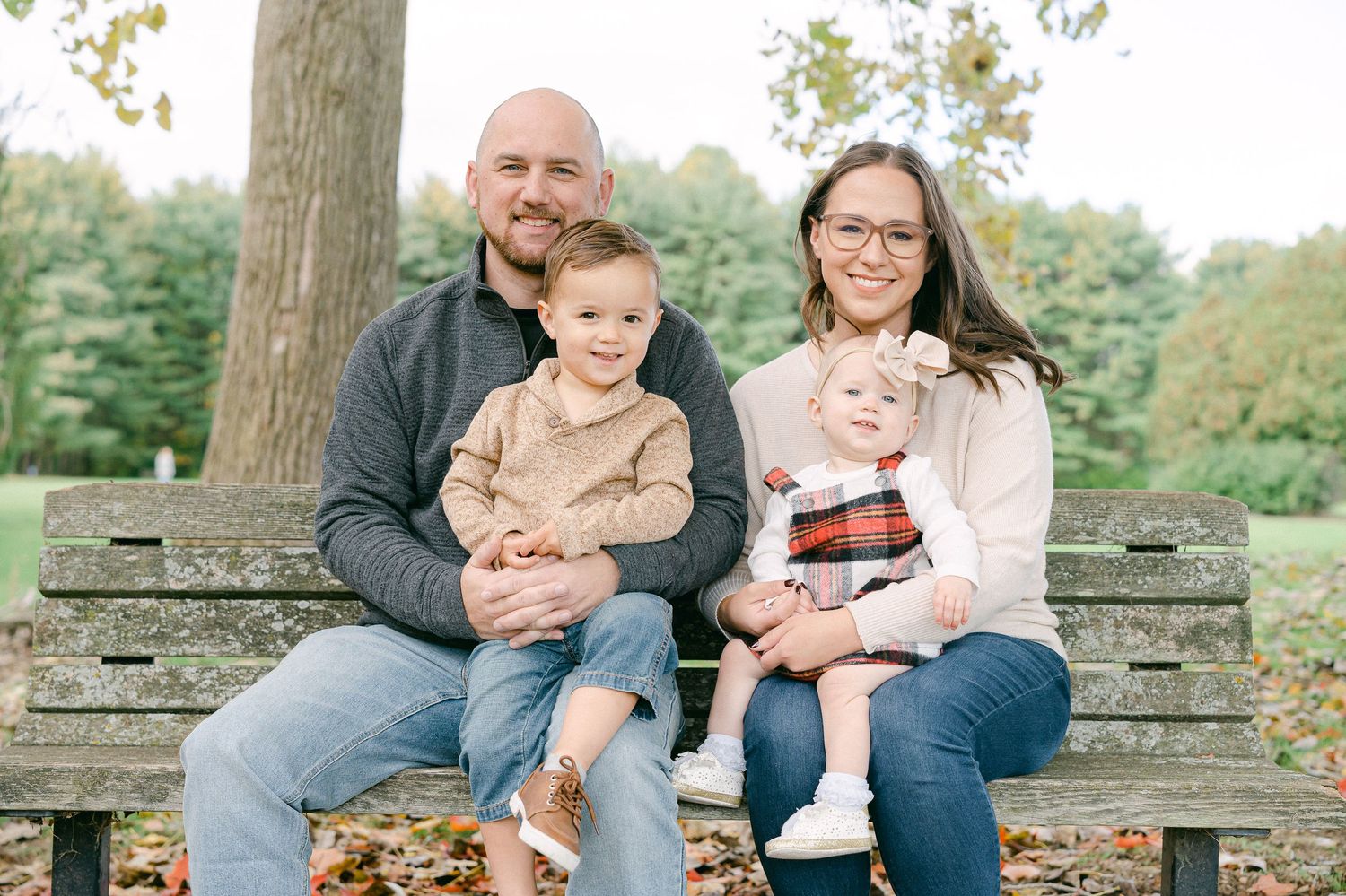 Family sits together on rustic wooden bench in autumn park setting during outdoor photo session.