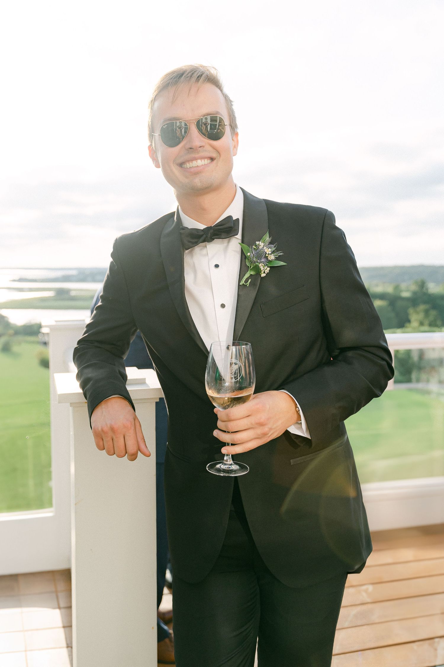 Smiling groom in black tuxedo and sunglasses holding champagne glass on outdoor balcony with scenic view in Cape Cod