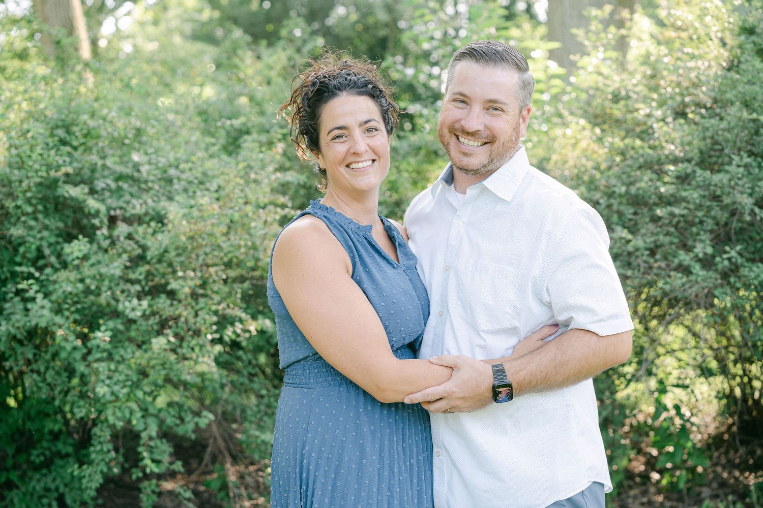 A couple embraces and smiles while standing together in a lush green garden setting on a sunny day.