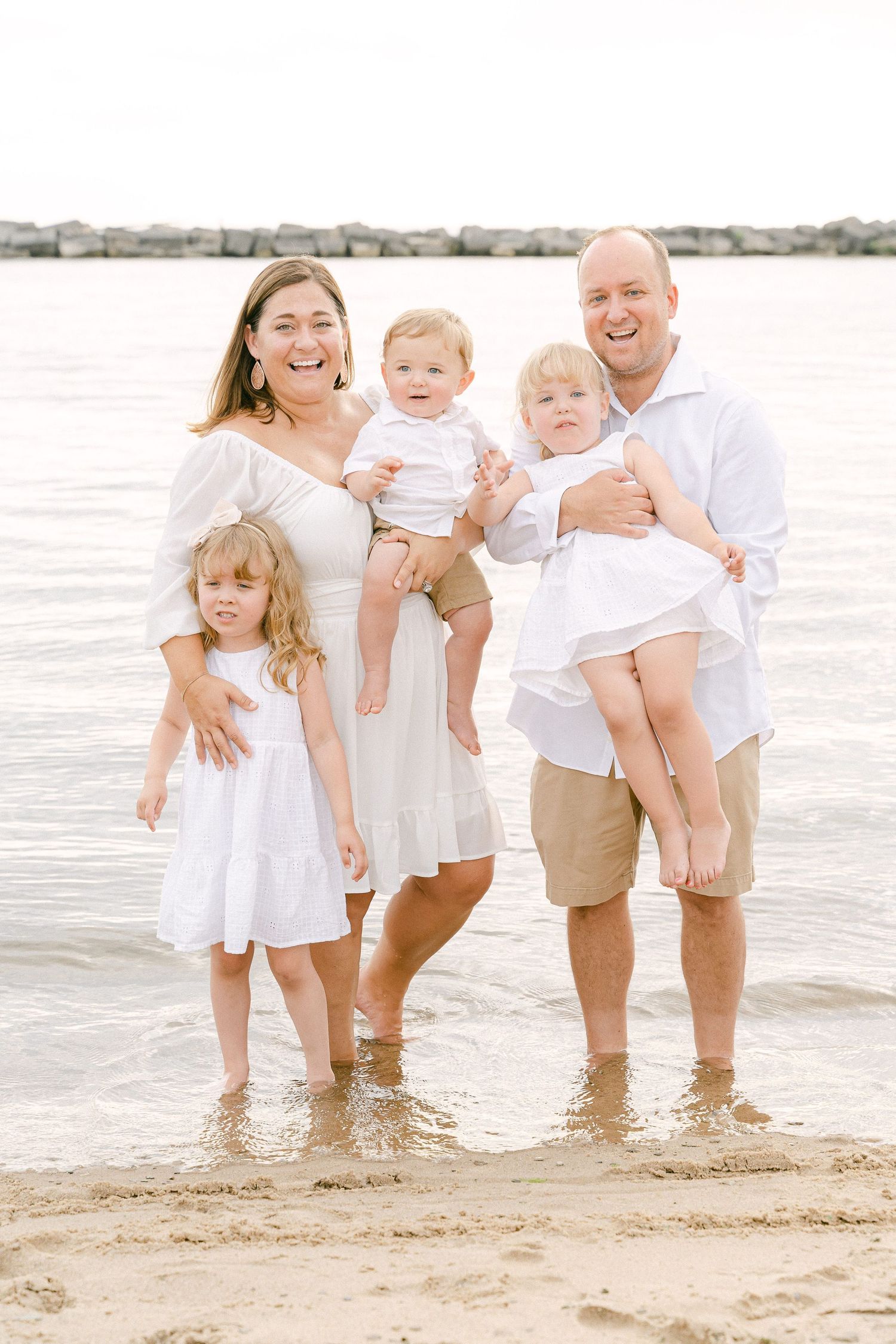 Family with young children playing together at the beach's edge in coordinated white outfits.