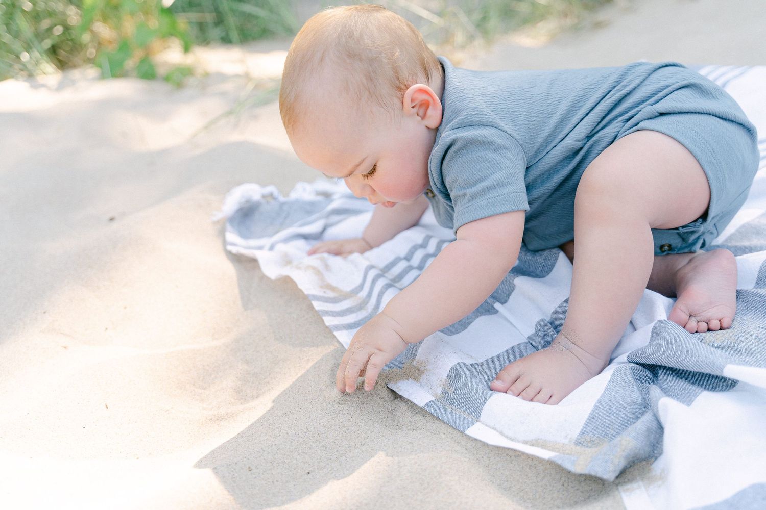 Baby in blue outfit crawls on striped blanket outdoors in soft sunlight.
