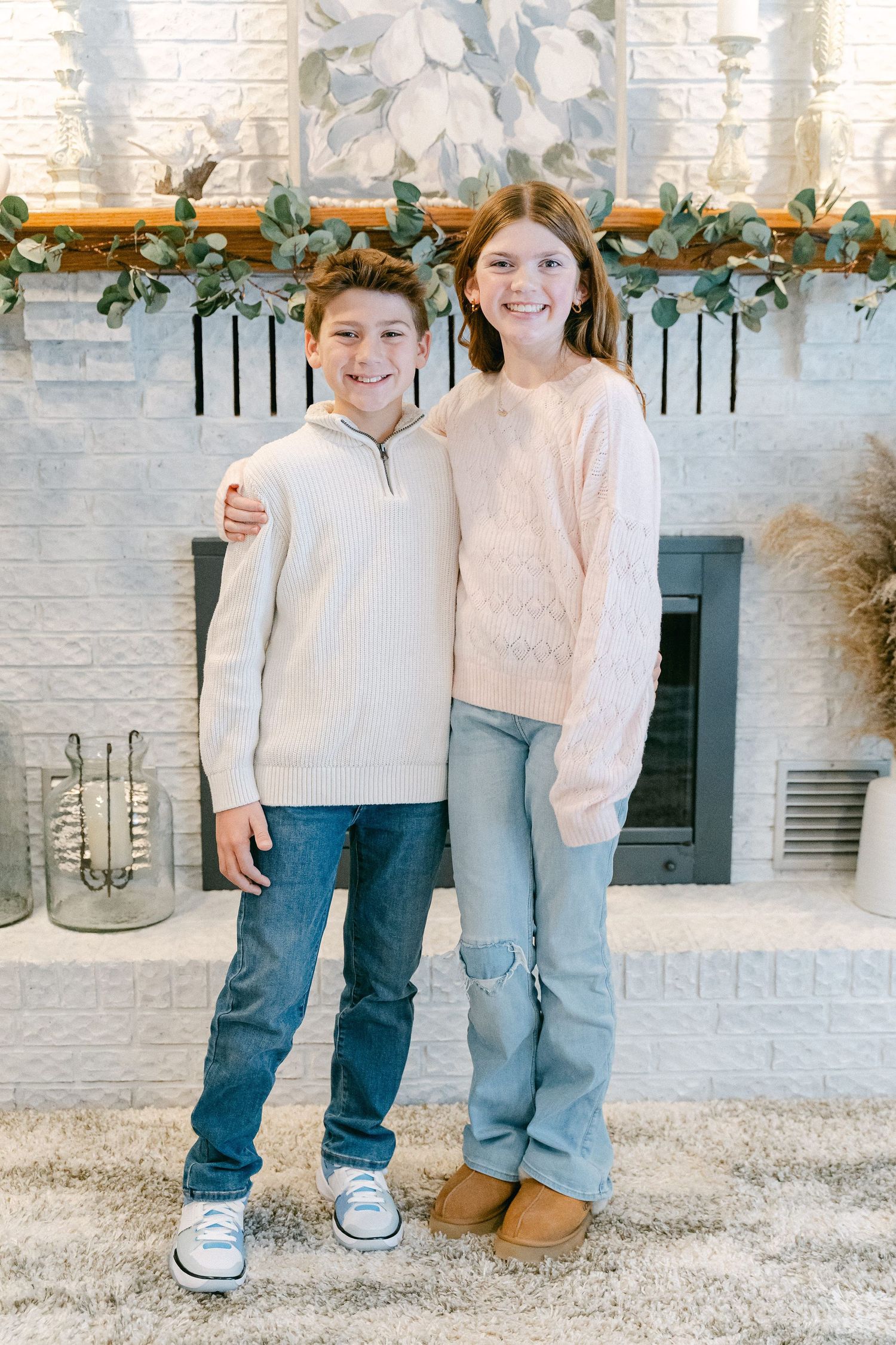 Children standing together in casual winter outfits against a white brick wall with holiday decor.