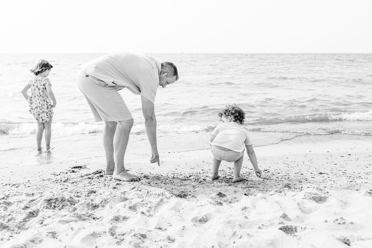Black and white photo of people exploring and playing together on a sandy beach by the ocean waves.