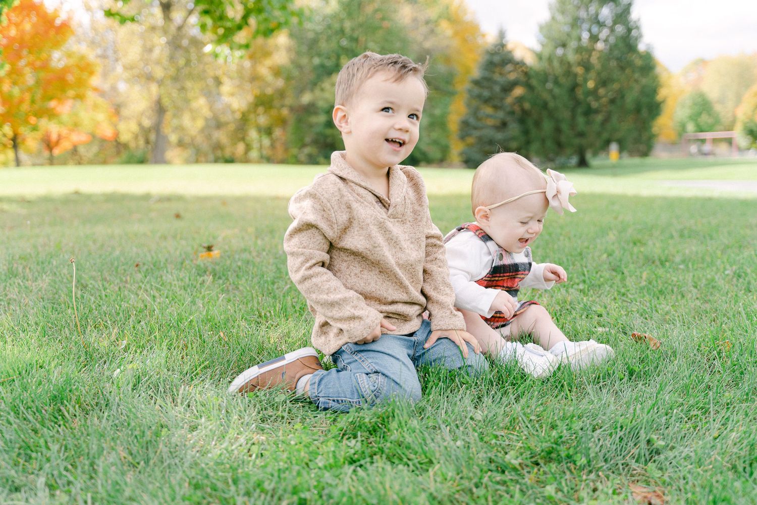 Two children sit together on grass in a park during autumn with fall foliage in the background.