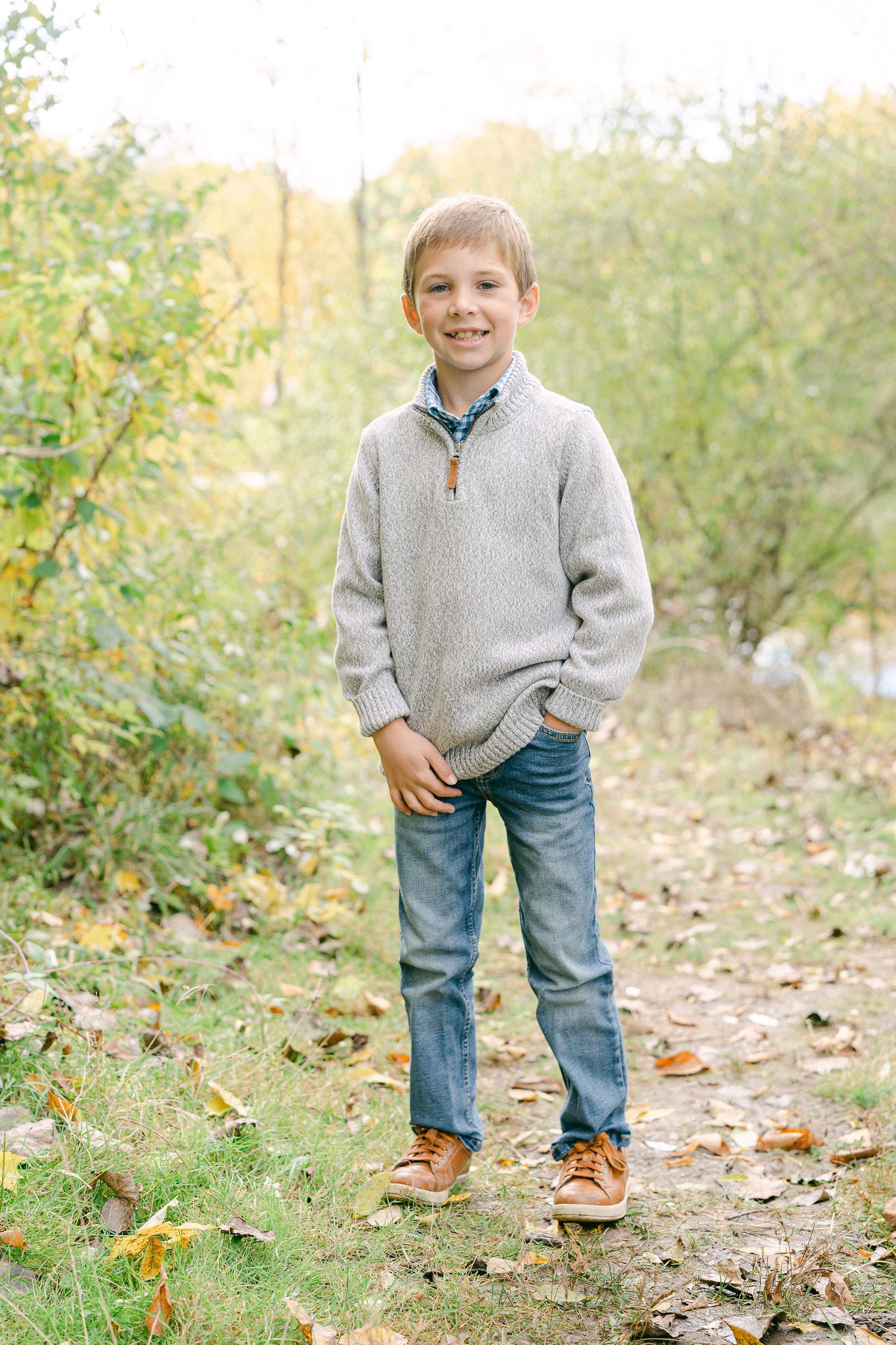 A small figure wearing a gray sweater and jeans stands on a leaf-covered path in an autumn outdoor setting.