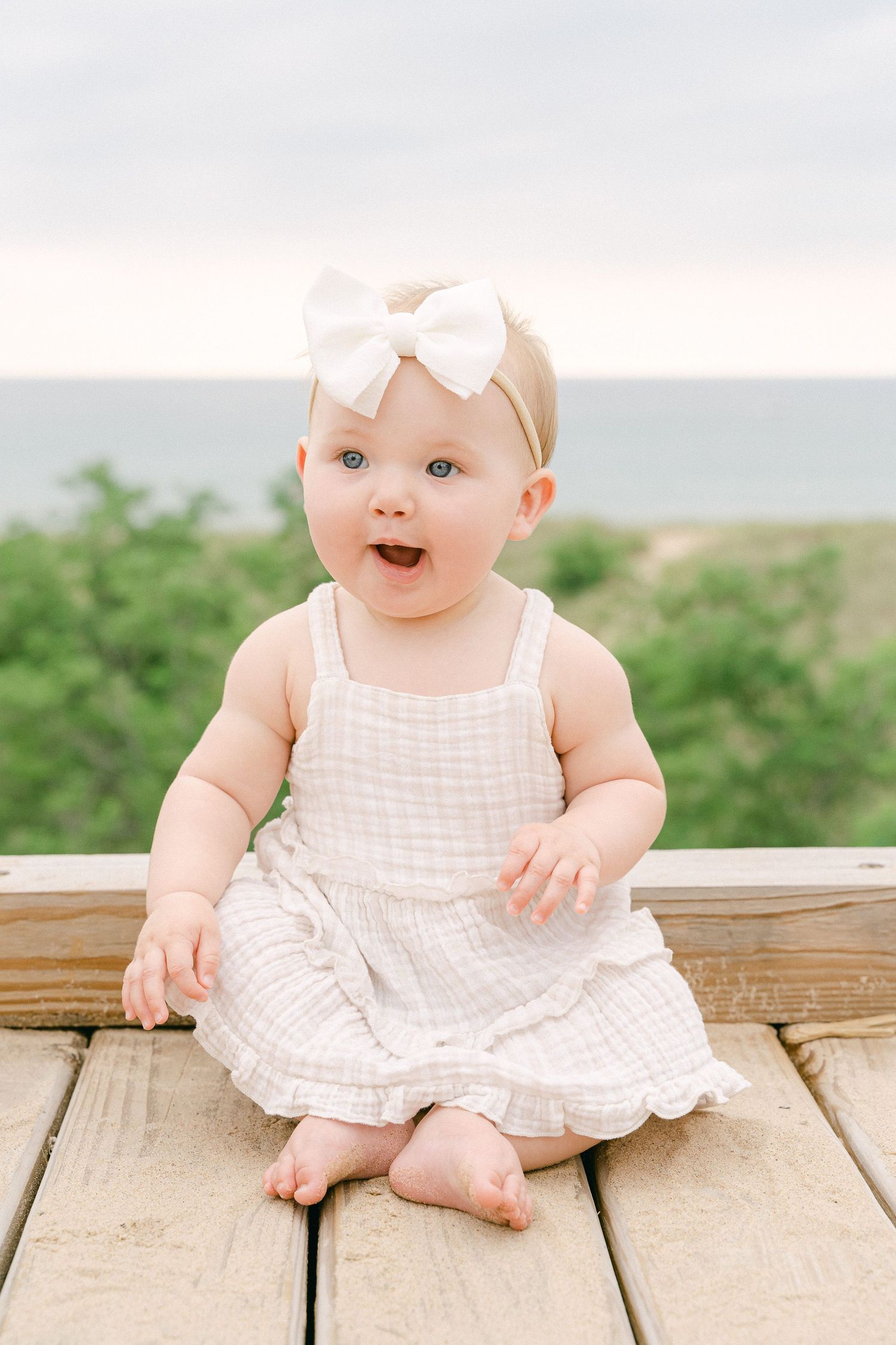 Baby in white ruffled dress sits on wooden deck outdoors with joyful expression.