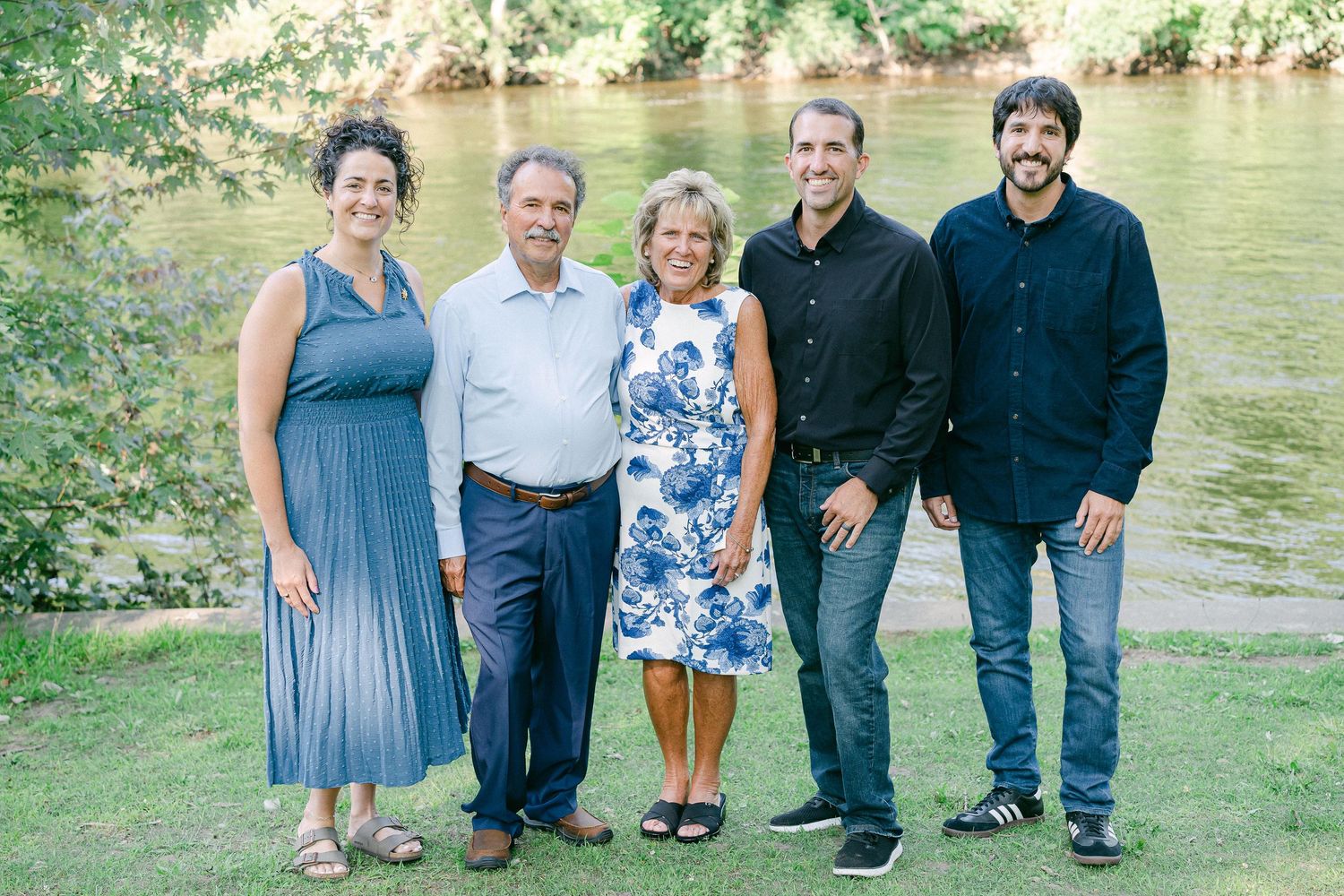 A group of people wearing casual attire pose together outdoors near a river on a sunny day.