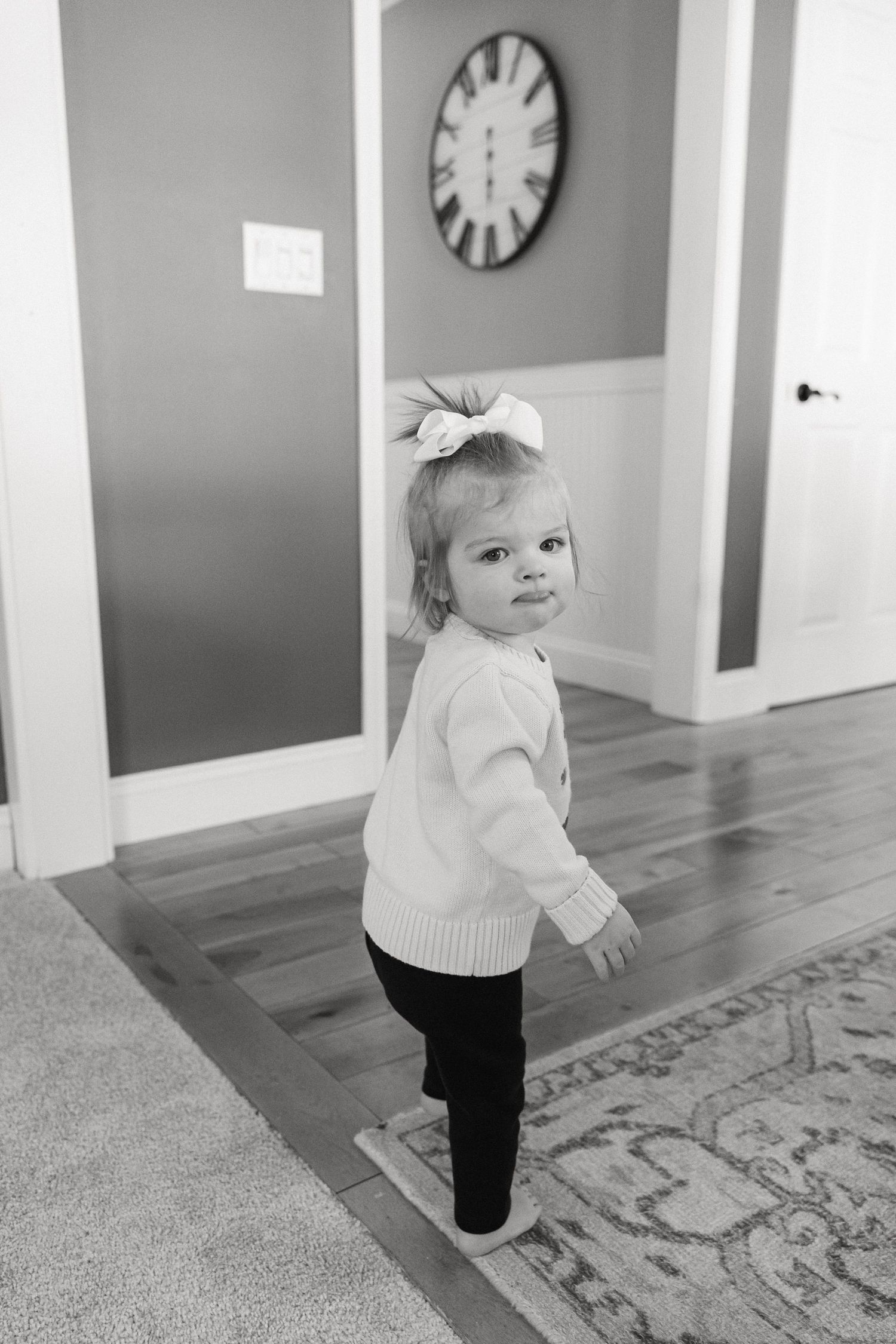 A series of three black and white photos showing a small toddler in a white sweater walking on hardwood floors and carpet.