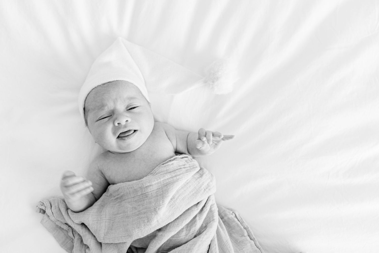 High key black and white image of a happy baby lying on white bedding.
