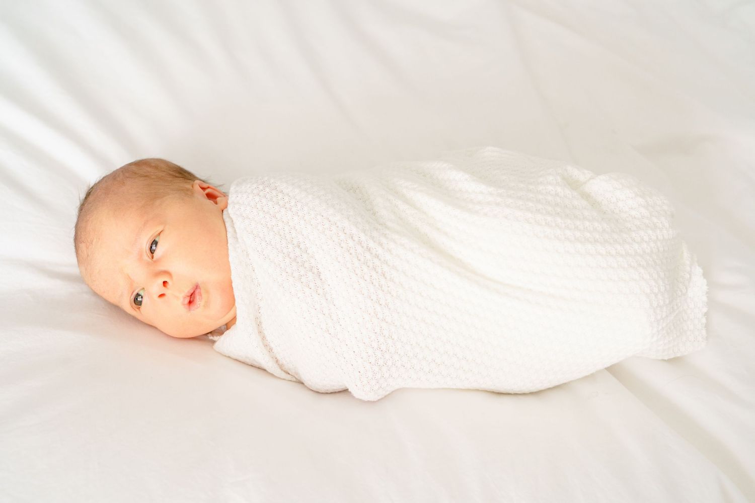 Alert baby swaddled in white blanket lying on soft white bedding.