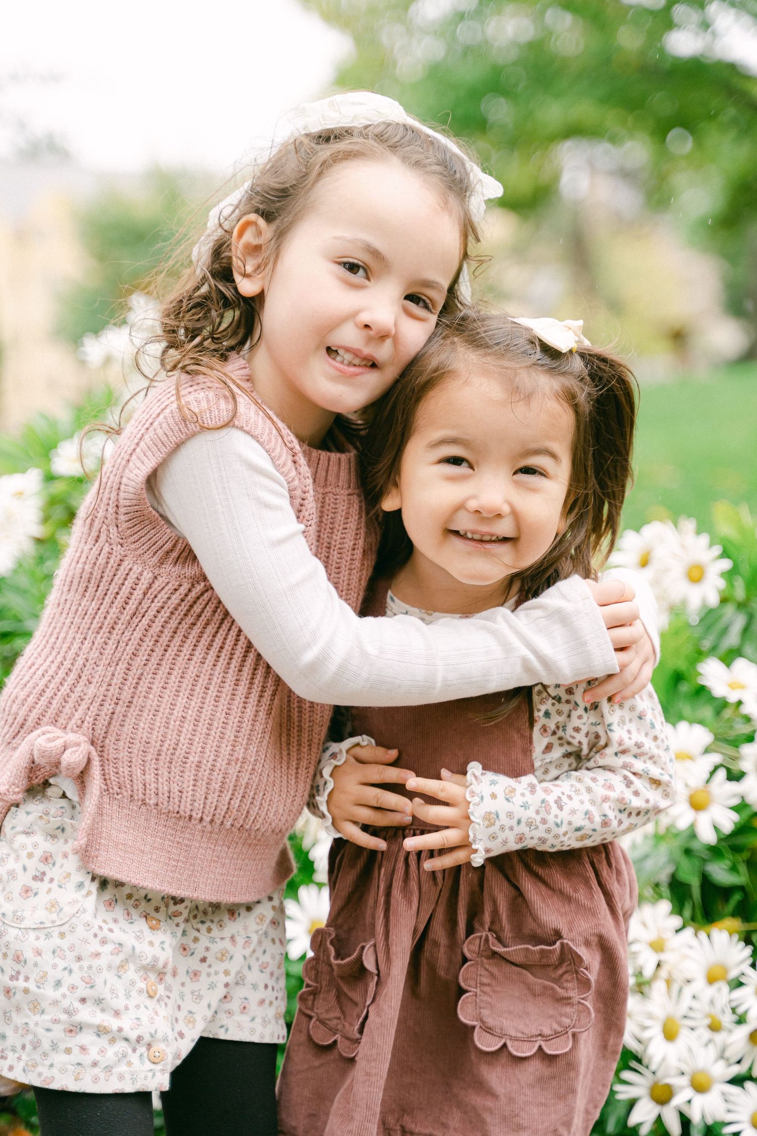 Two children in pink and brown outfits play together among blooming white daisies in a garden setting.