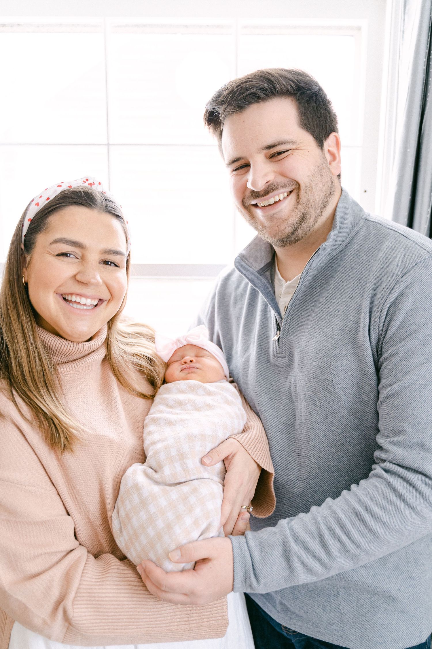 A couple in pink and grey sweaters share joyful moments with their newborn baby in a bright indoor setting.