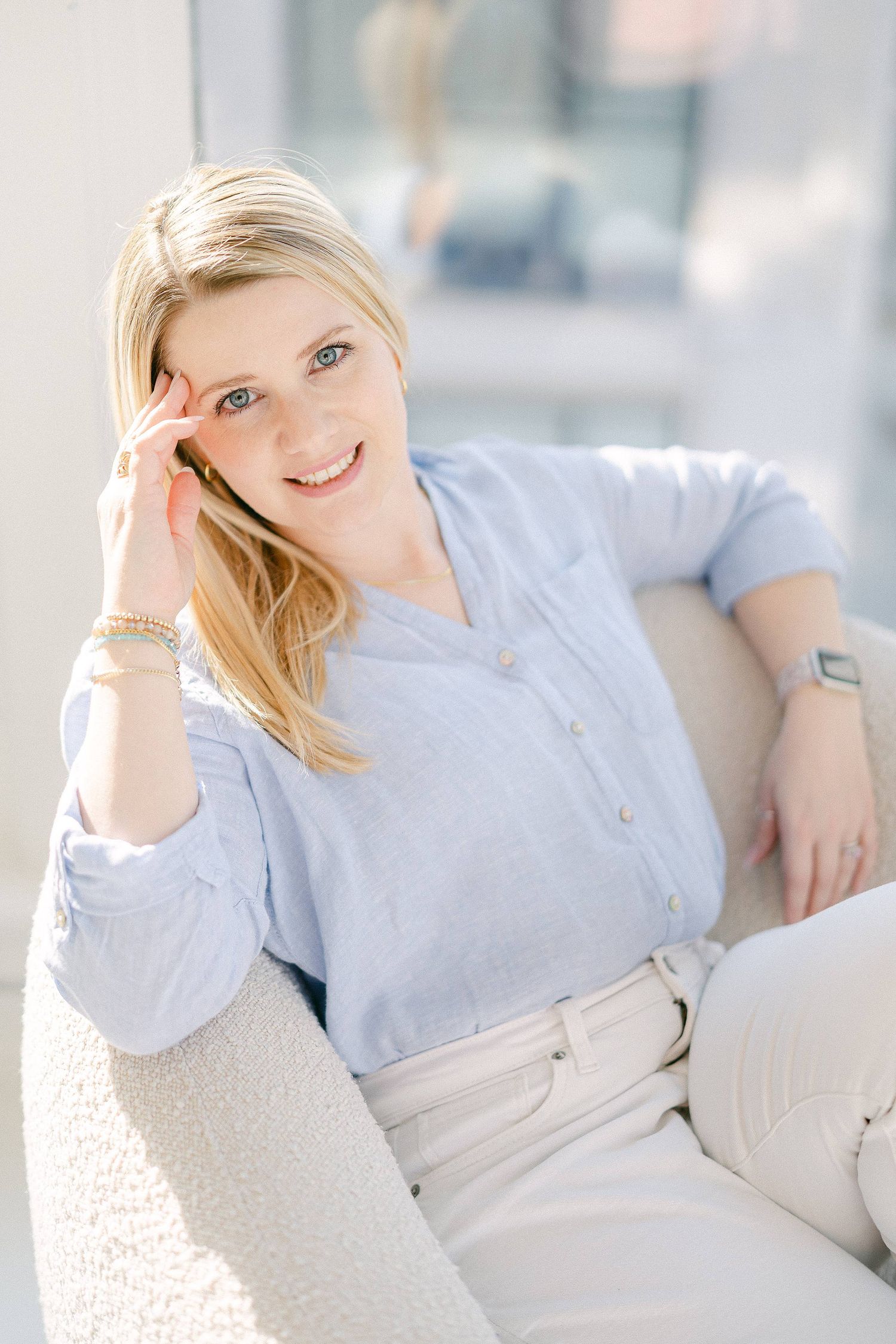 Woman in light blue shirt relaxes on white couch in sunlit room.