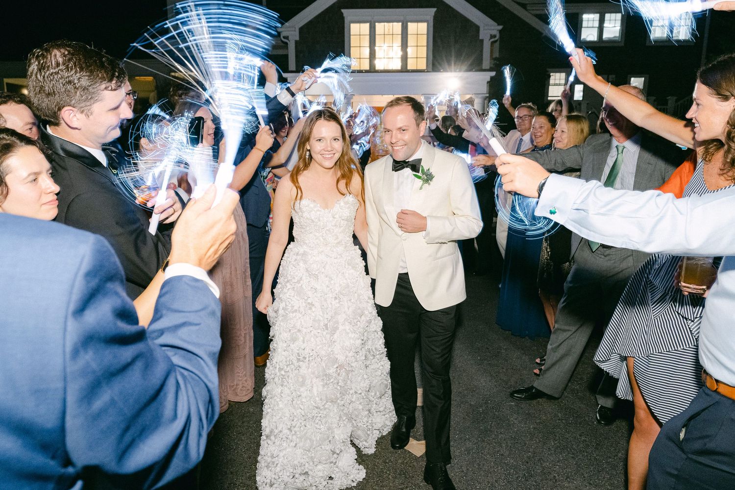 Bride and groom exit wedding reception through sparkler send-off tunnel surrounded by cheering guests at night in Cape Cod