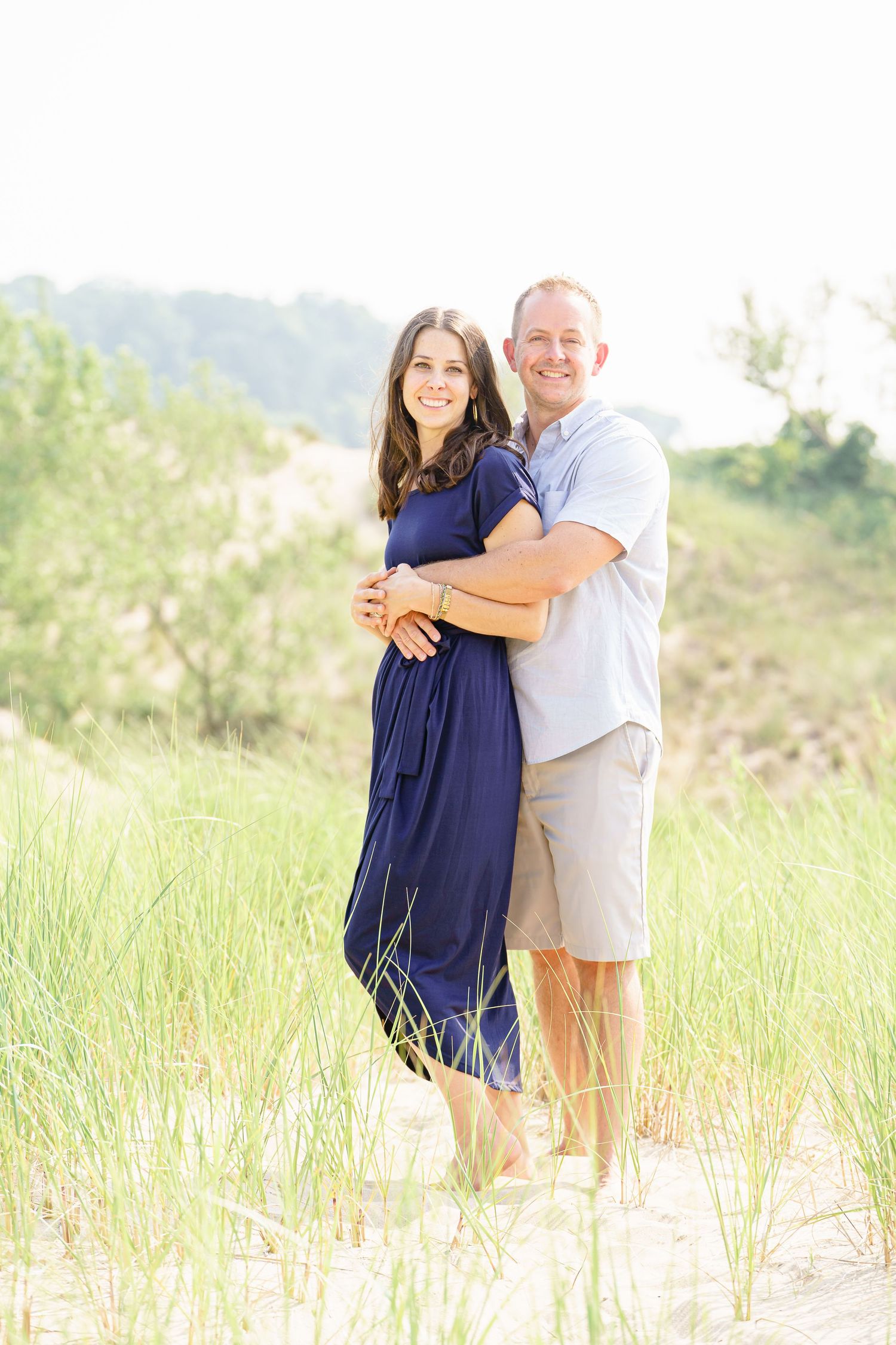 A couple embraces lovingly on a sandy beach with tall grass and soft sunlight creating a romantic atmosphere.