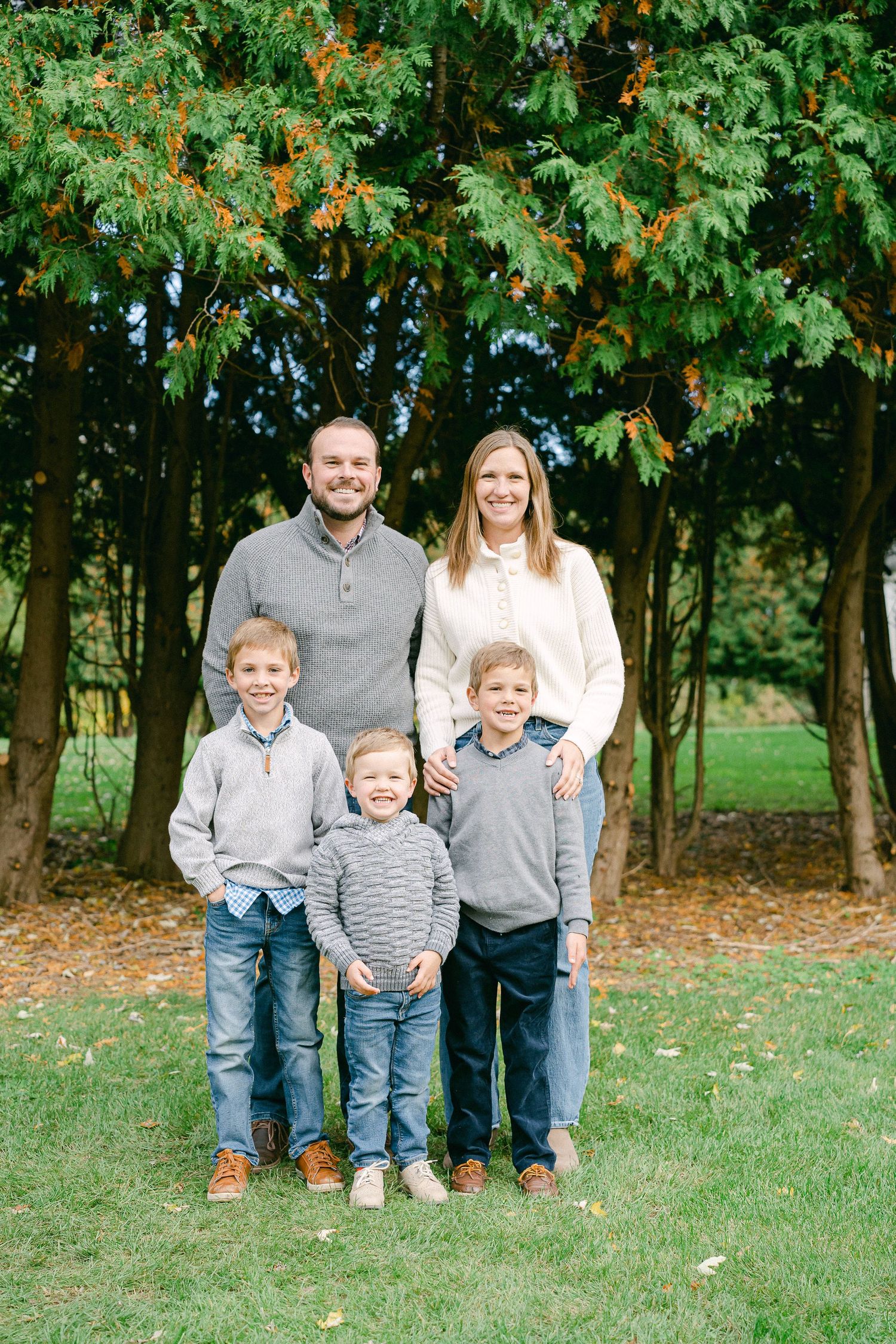 A group poses together outdoors on grass with autumn trees in the background wearing coordinated gray and white outfits.