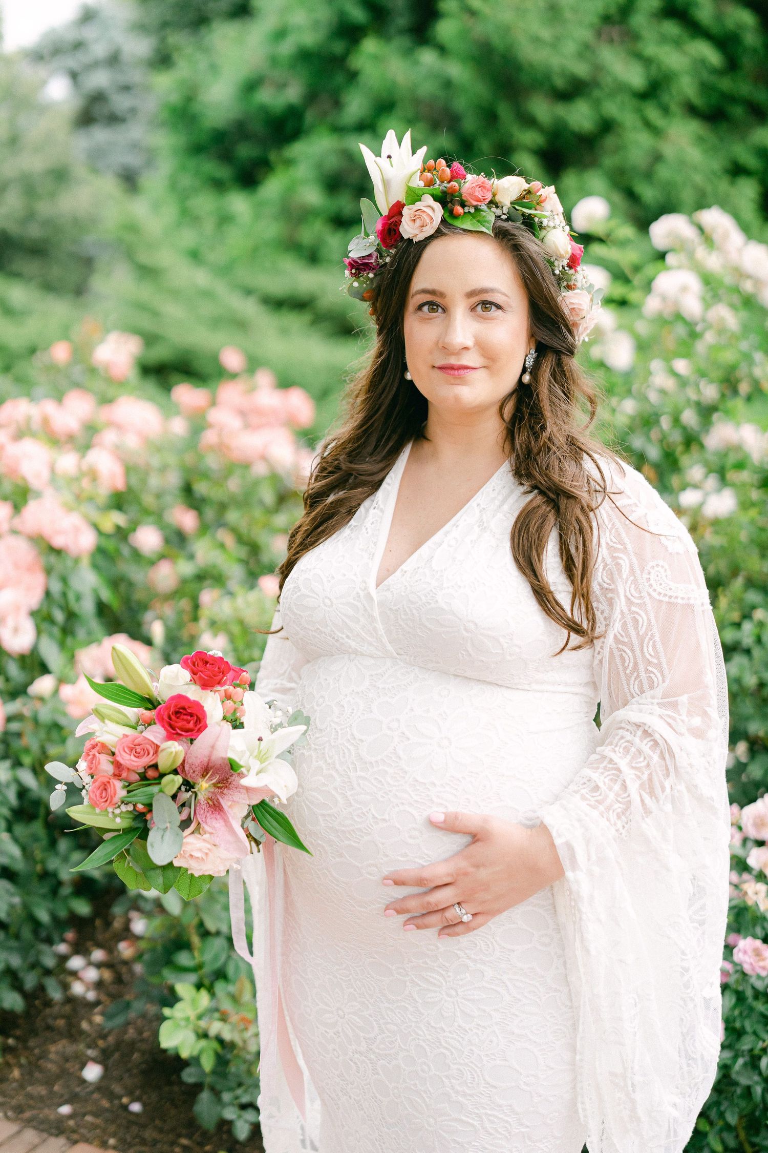 A pregnant woman in a white lace dress stands in a garden wearing a floral crown and holding a pink bouquet.