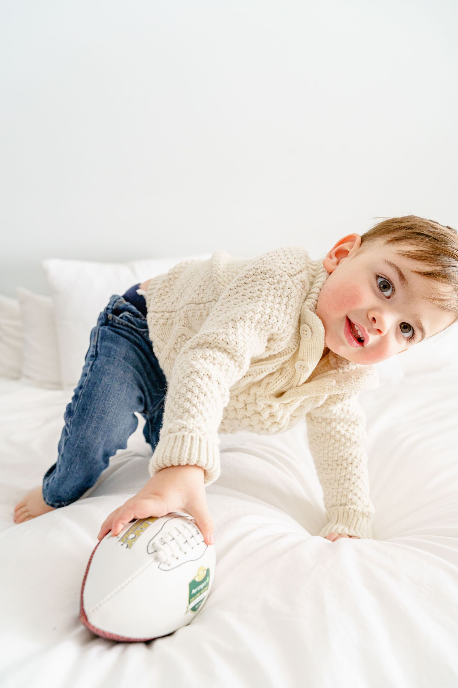 Child in cable knit sweater and jeans lying on white bedding with a rugby ball.