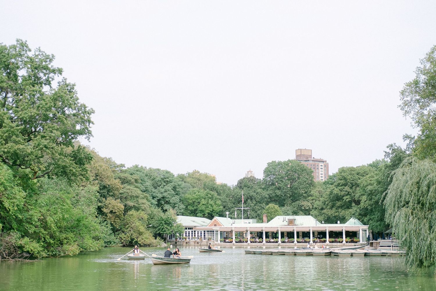 A scenic view of The Loeb Boathouse in Central Park with boats on the lake and lush greenery surrounding it.