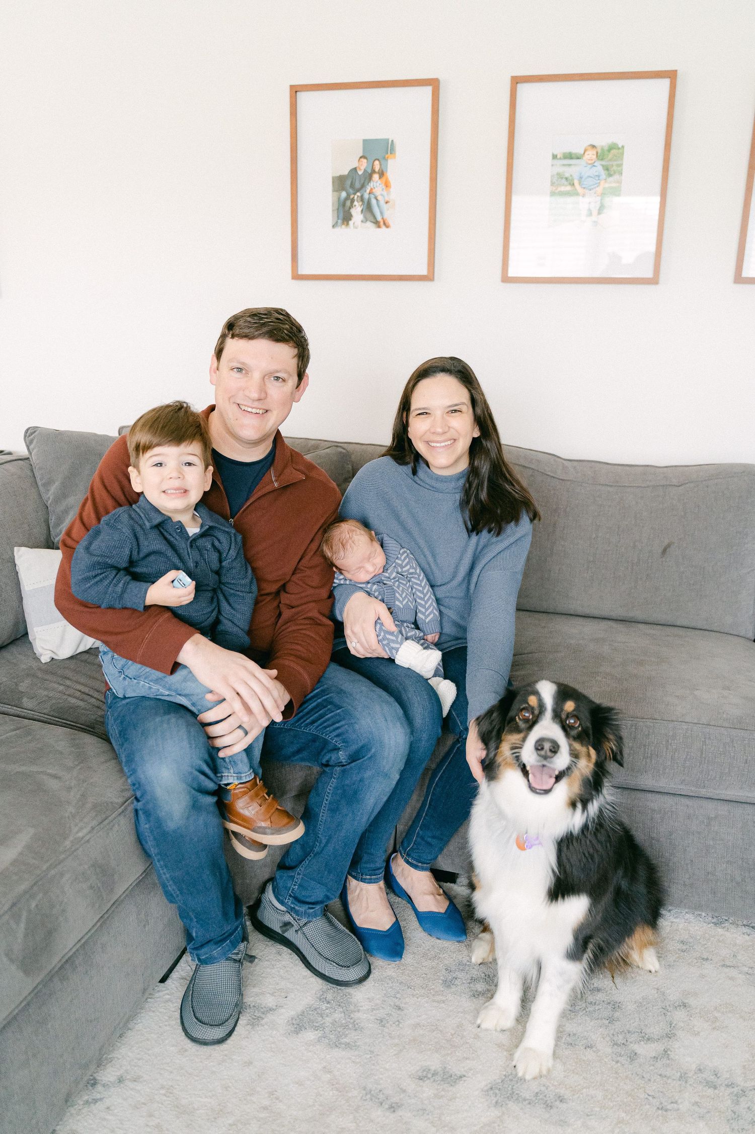 Family sitting together on a gray couch with their Australian Shepherd dog in a living room with framed art on the wall.