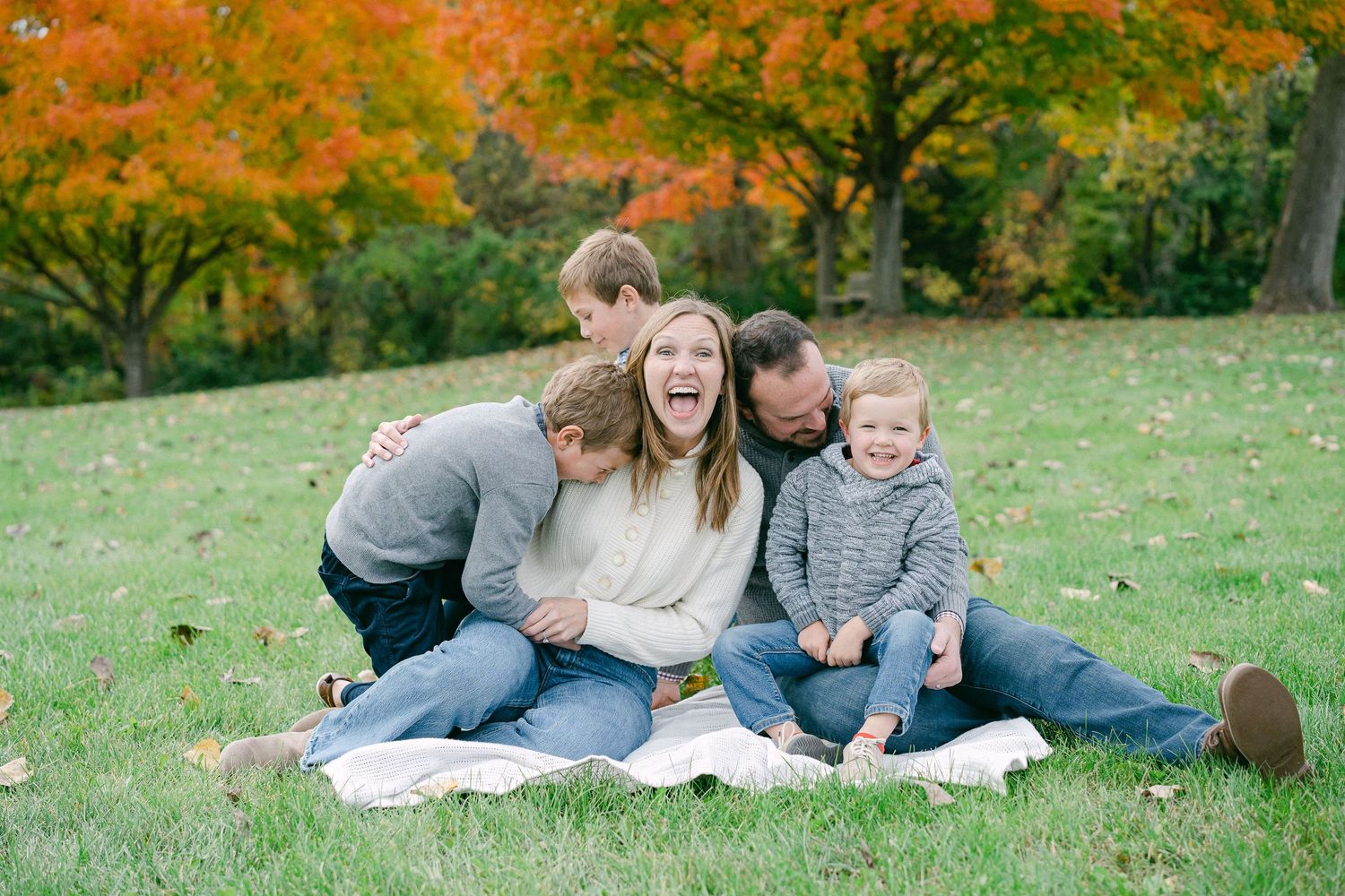 Family relaxes together on blanket during autumn photoshoot with colorful fall foliage in background.