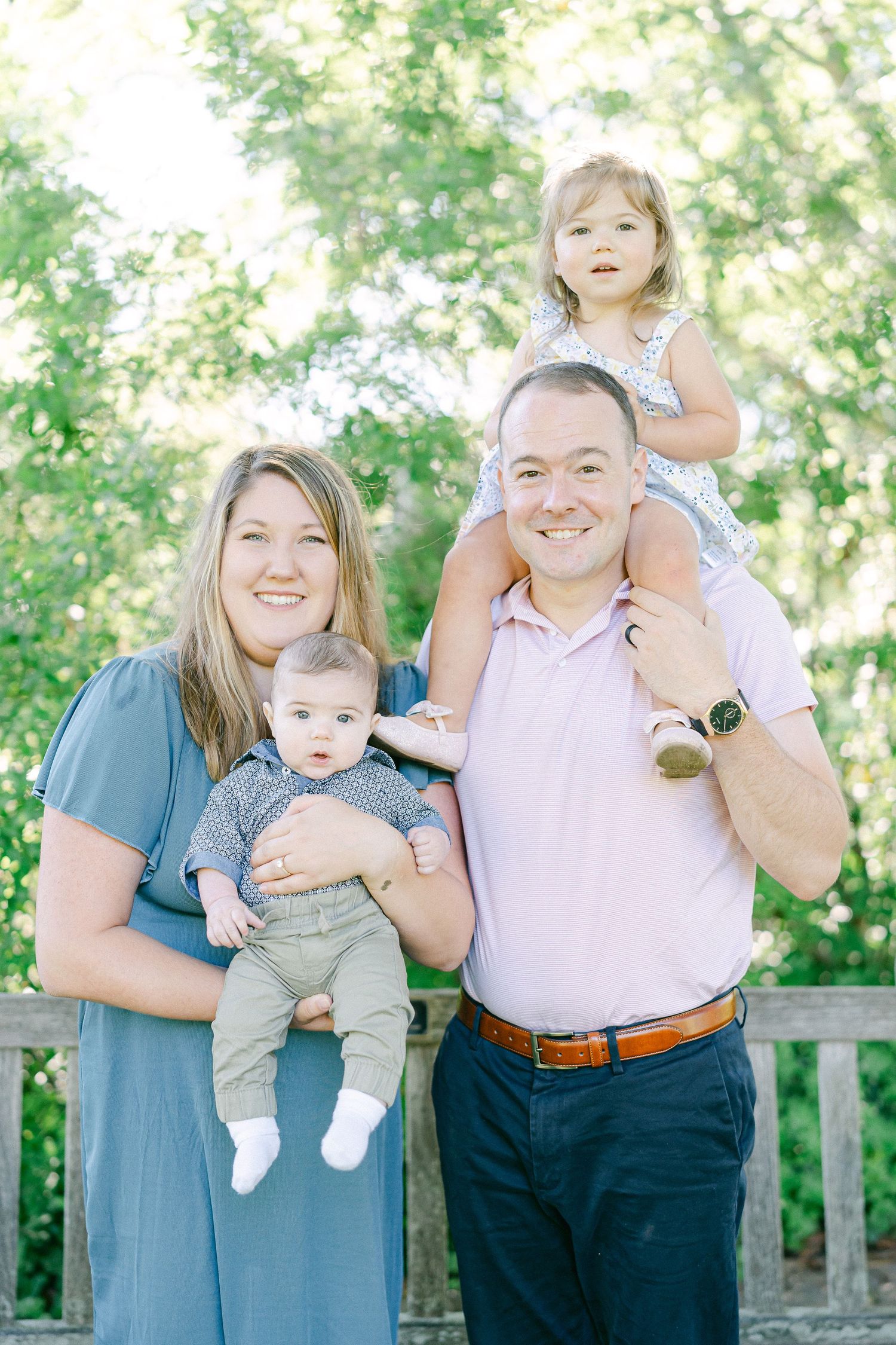 A happy family poses together outdoors on a sunny day with trees in the background.