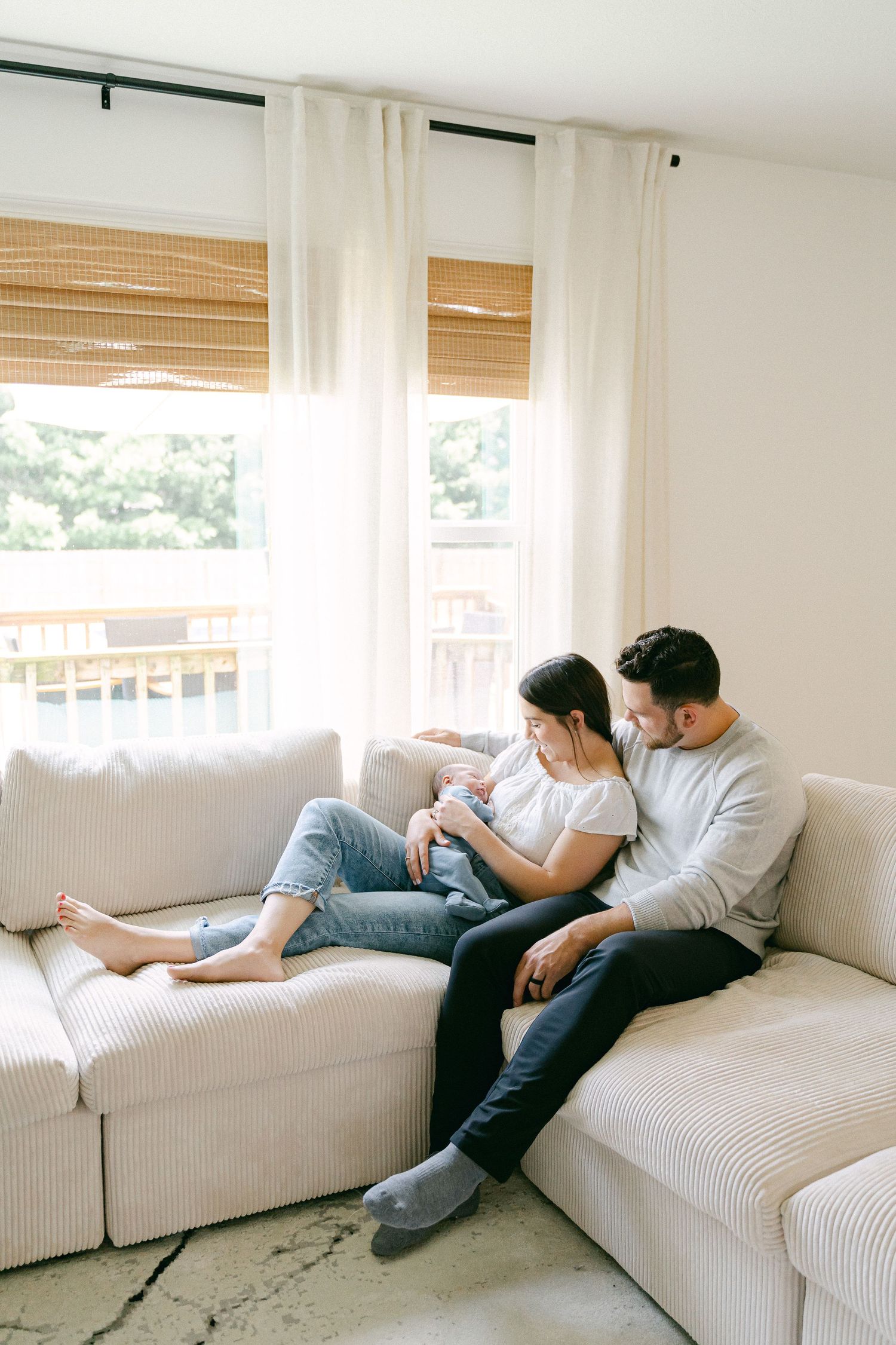 A couple sits together on a white sectional sofa near windows with sheer curtains and bamboo shades.
