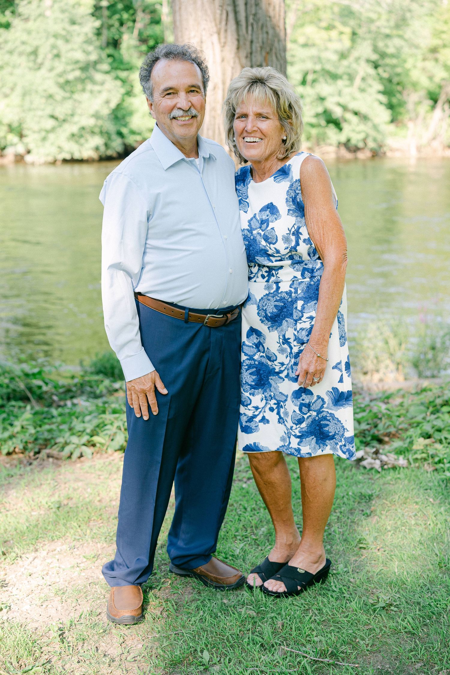 A couple in blue and white attire poses together by a lake on a sunny summer day.