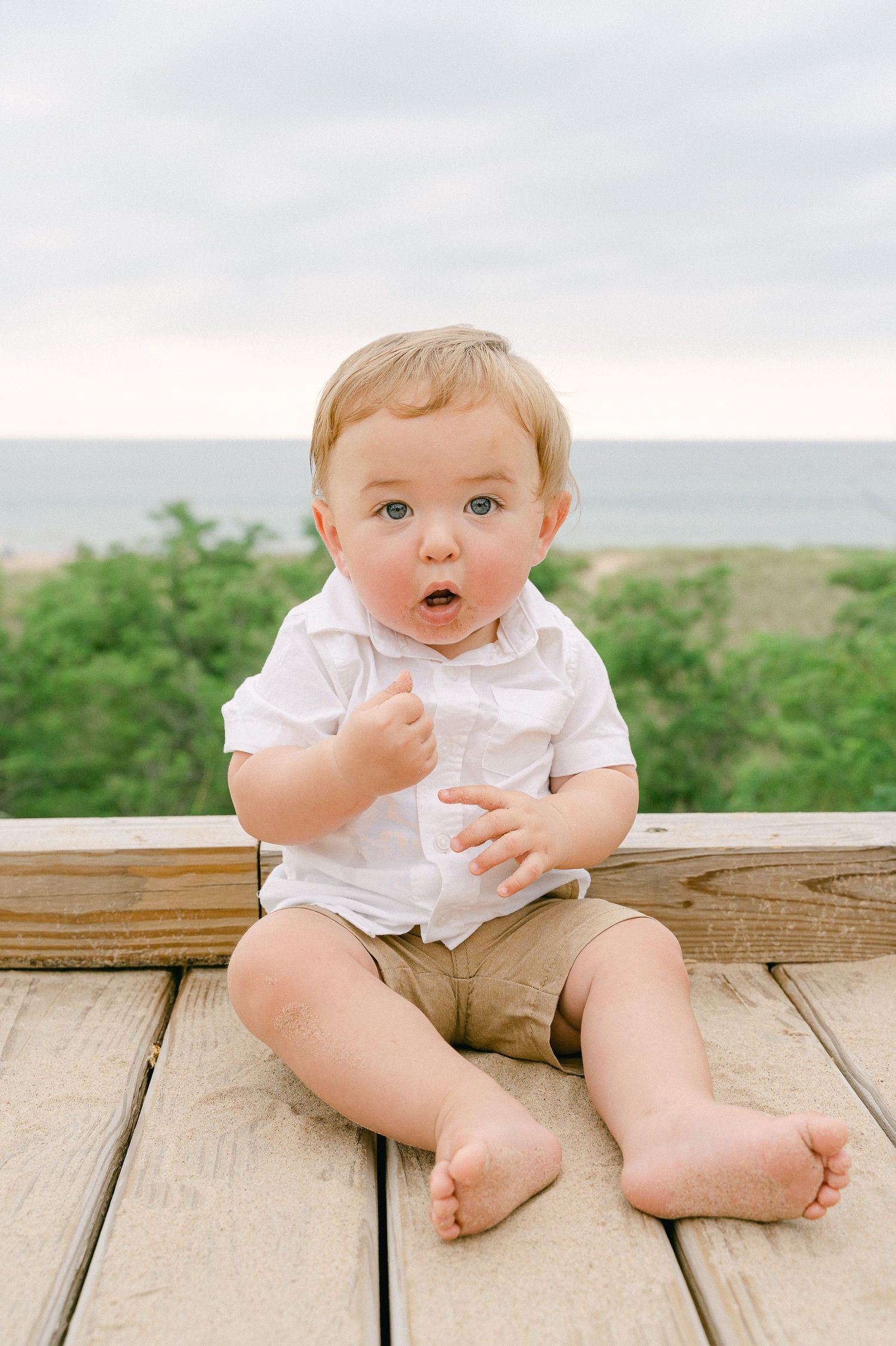 Baby in white outfit sits on wooden deck boards with natural scenery in background.