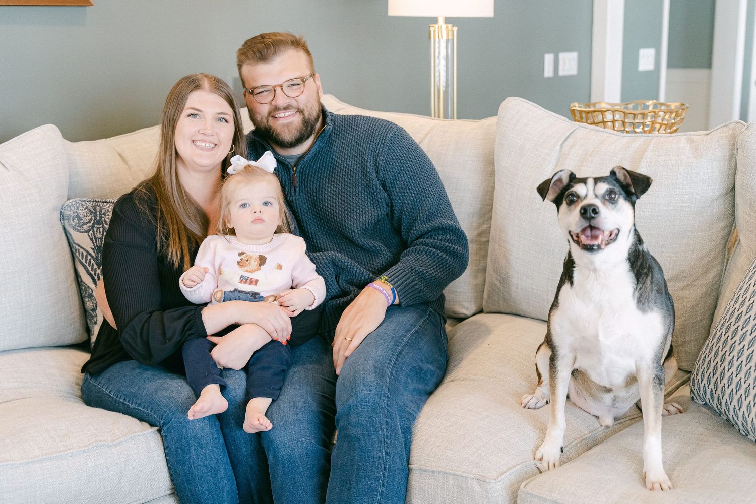 A family sits together on a gray couch with their black and white dog in a bright modern living room.