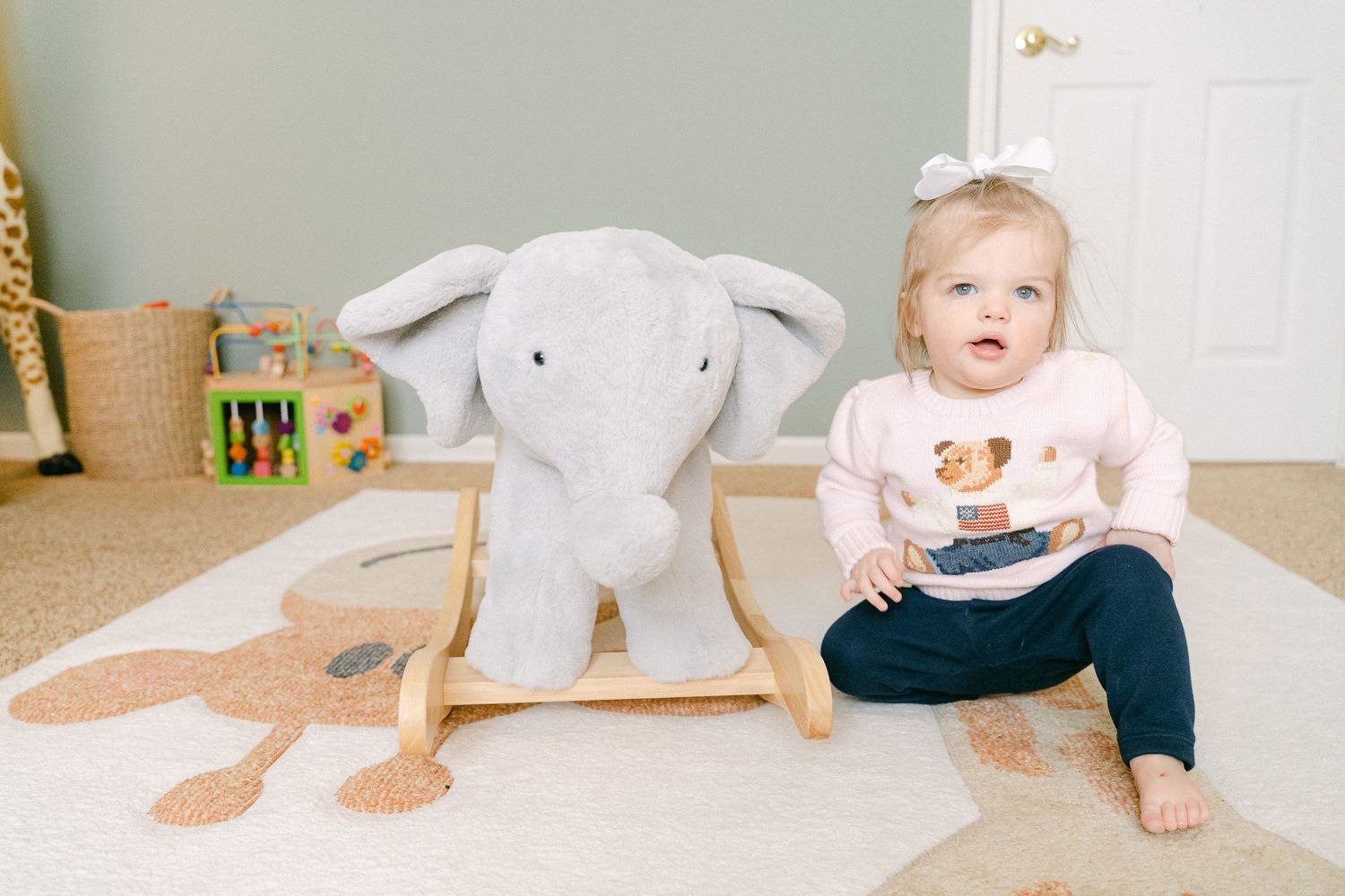 A baby in a white outfit crawls on a beige carpet near a stuffed elephant toy.