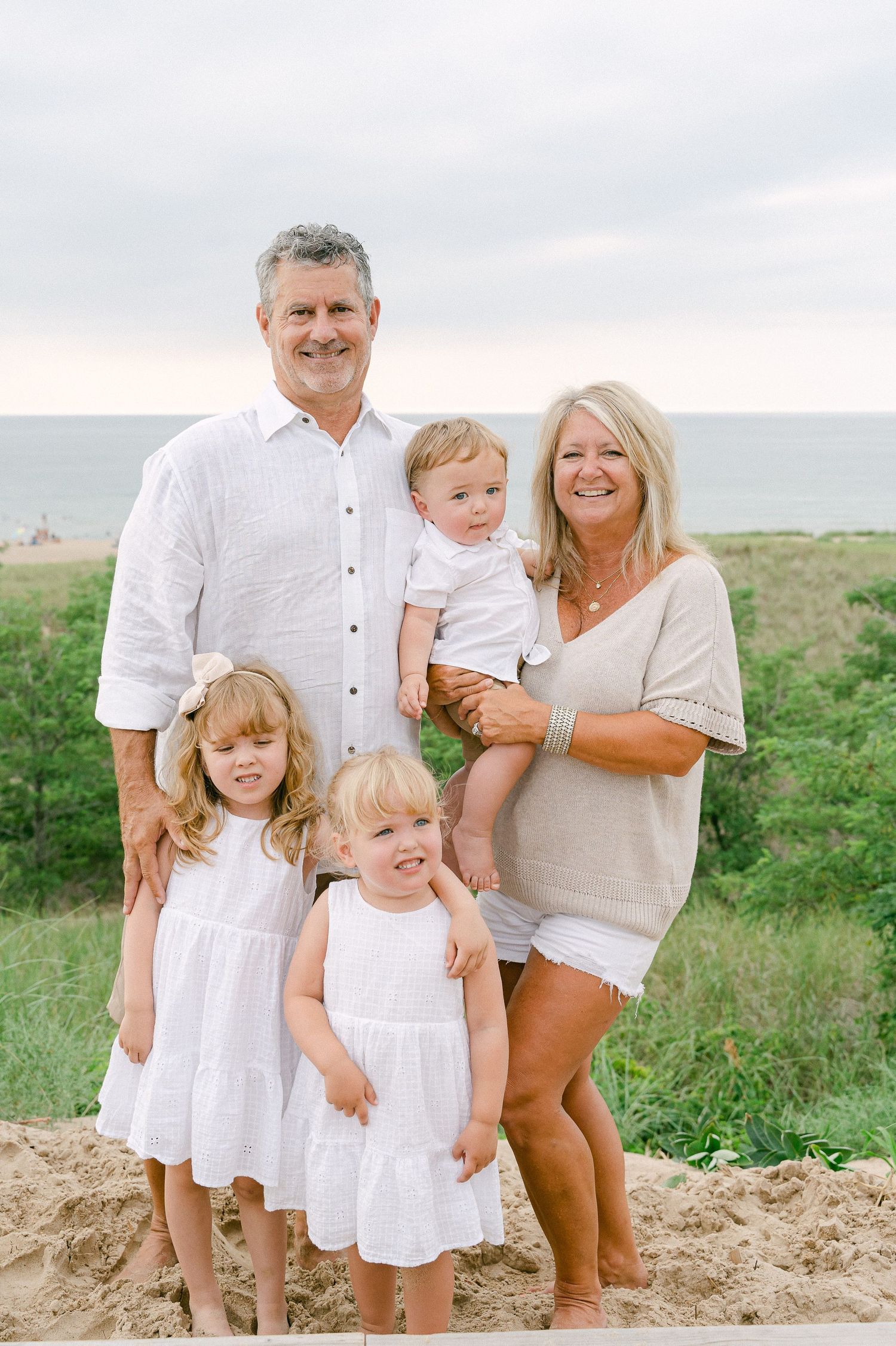 Family wearing coordinated white outfits poses together on sandy hill with green foliage in background.