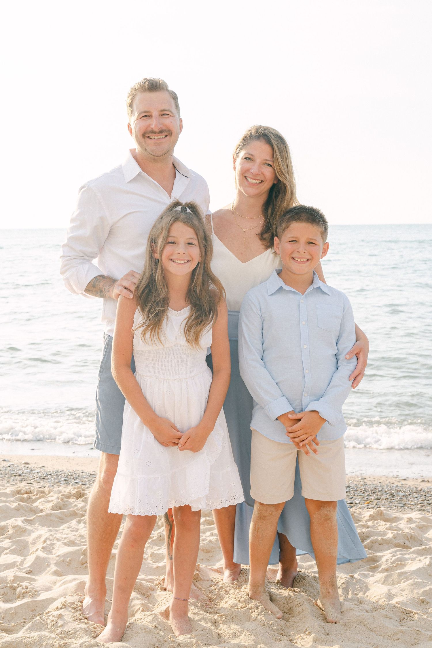 A group wearing coordinated white and light blue outfits poses together on a sunny beach with ocean waves in the background.