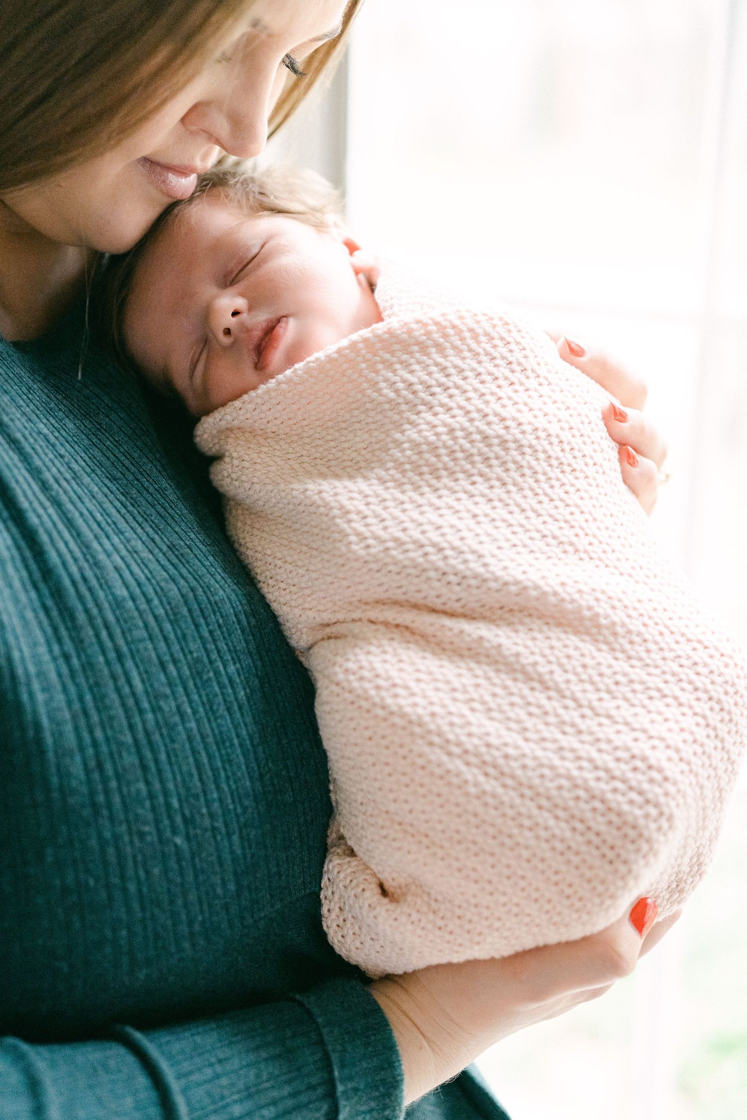 A sleeping newborn wrapped in a soft white knit blanket is held close against a teal sweater.