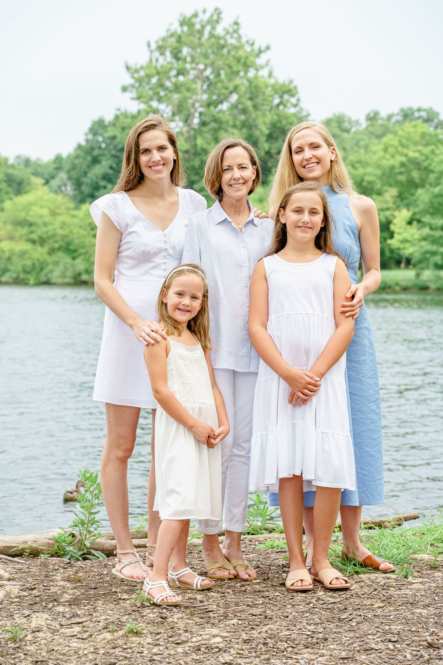 A group in light summer dresses poses together by a serene lake on a sunny day.