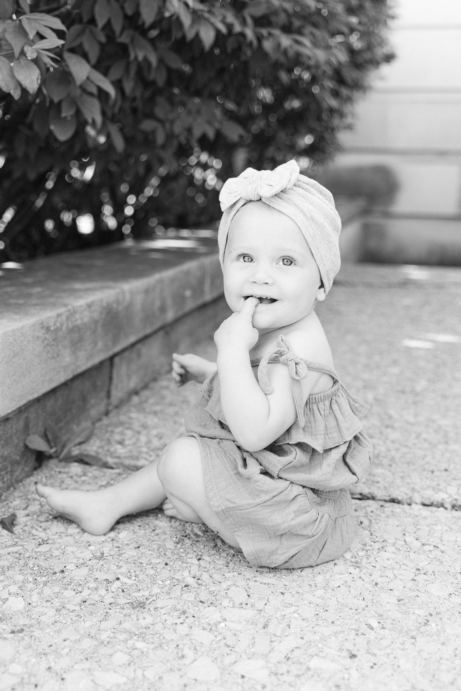 Black and white photo of a baby in a turban bow headband sitting on concrete with finger in mouth.