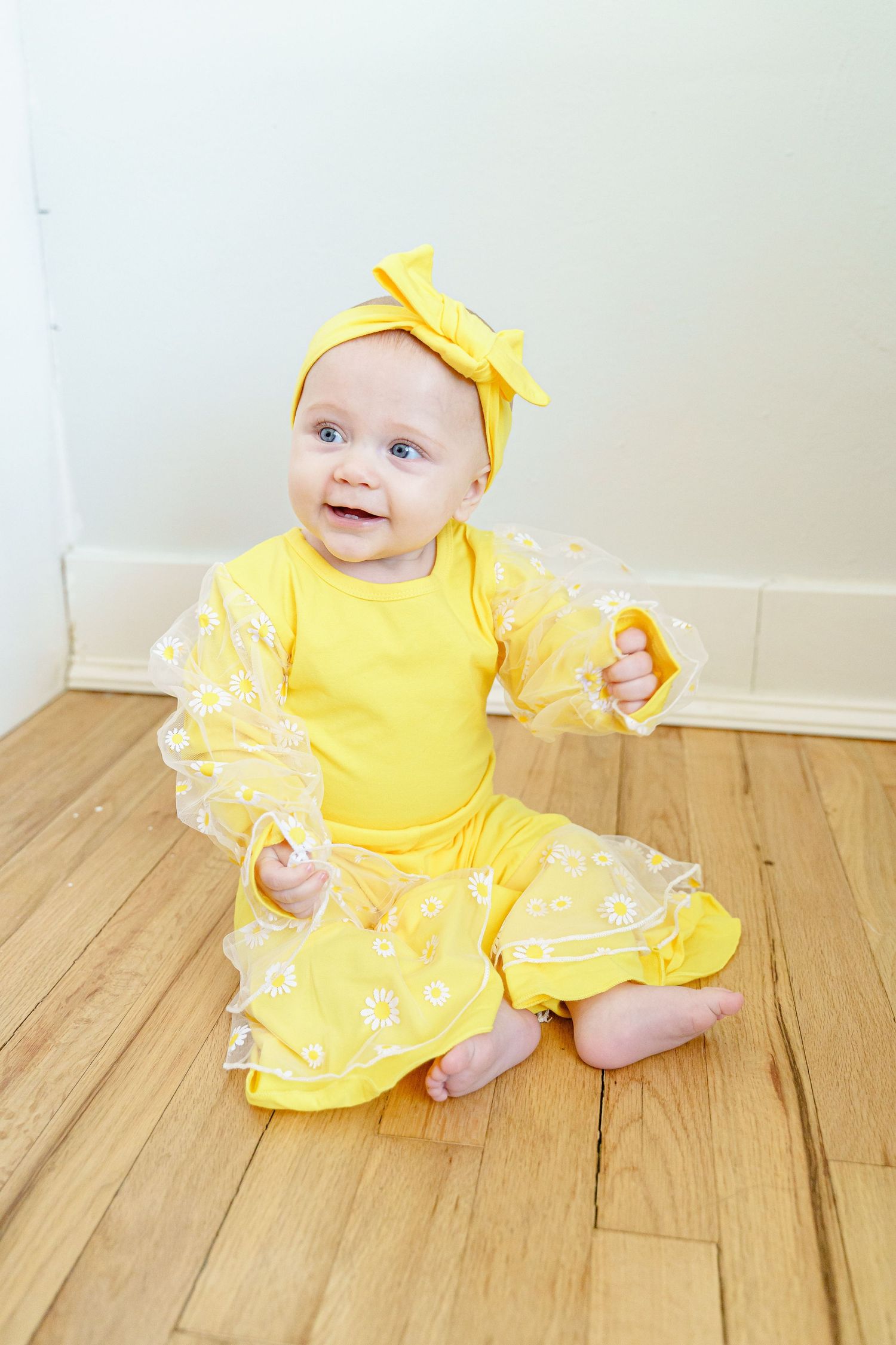 A baby in a yellow dress with lace details sits on hardwood flooring.