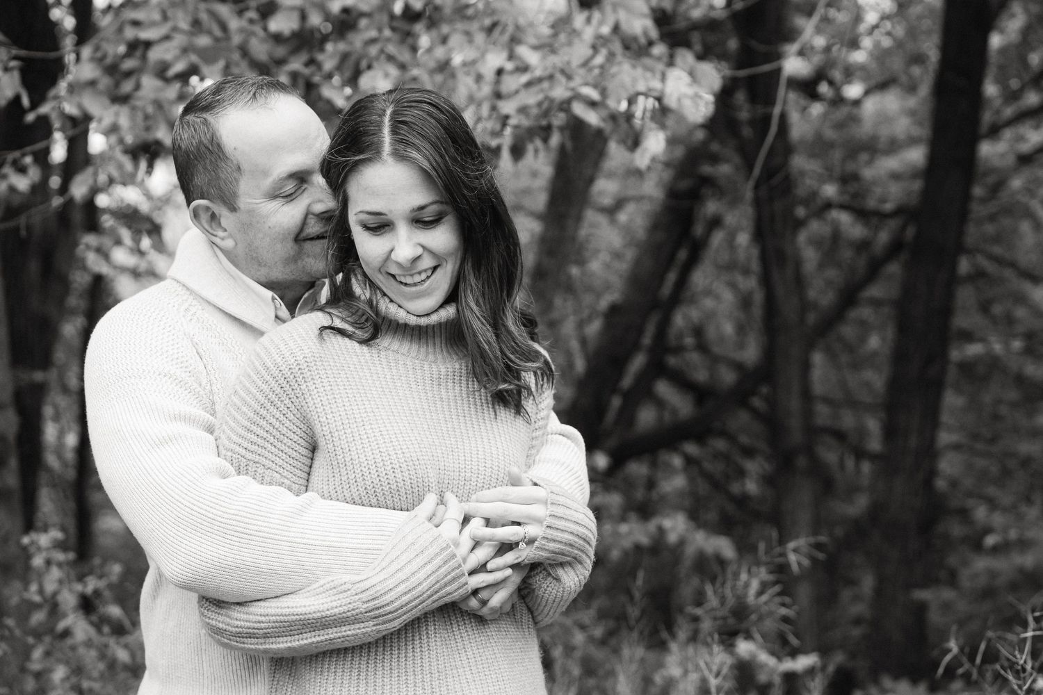 Black and white portrait of family members embracing and smiling together outdoors among trees.
