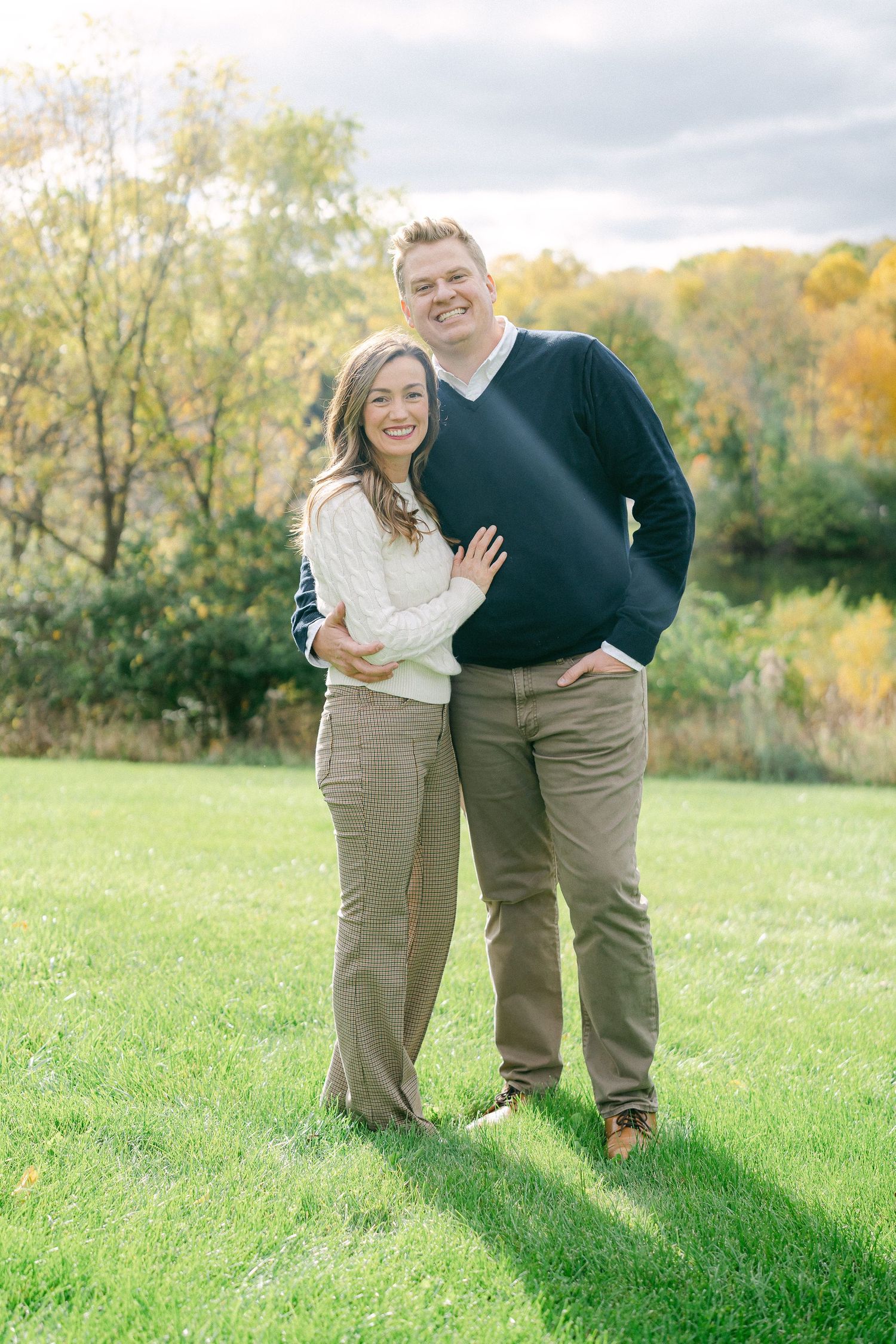 A couple stands together on grass with fall foliage in the background wearing coordinated sweaters.