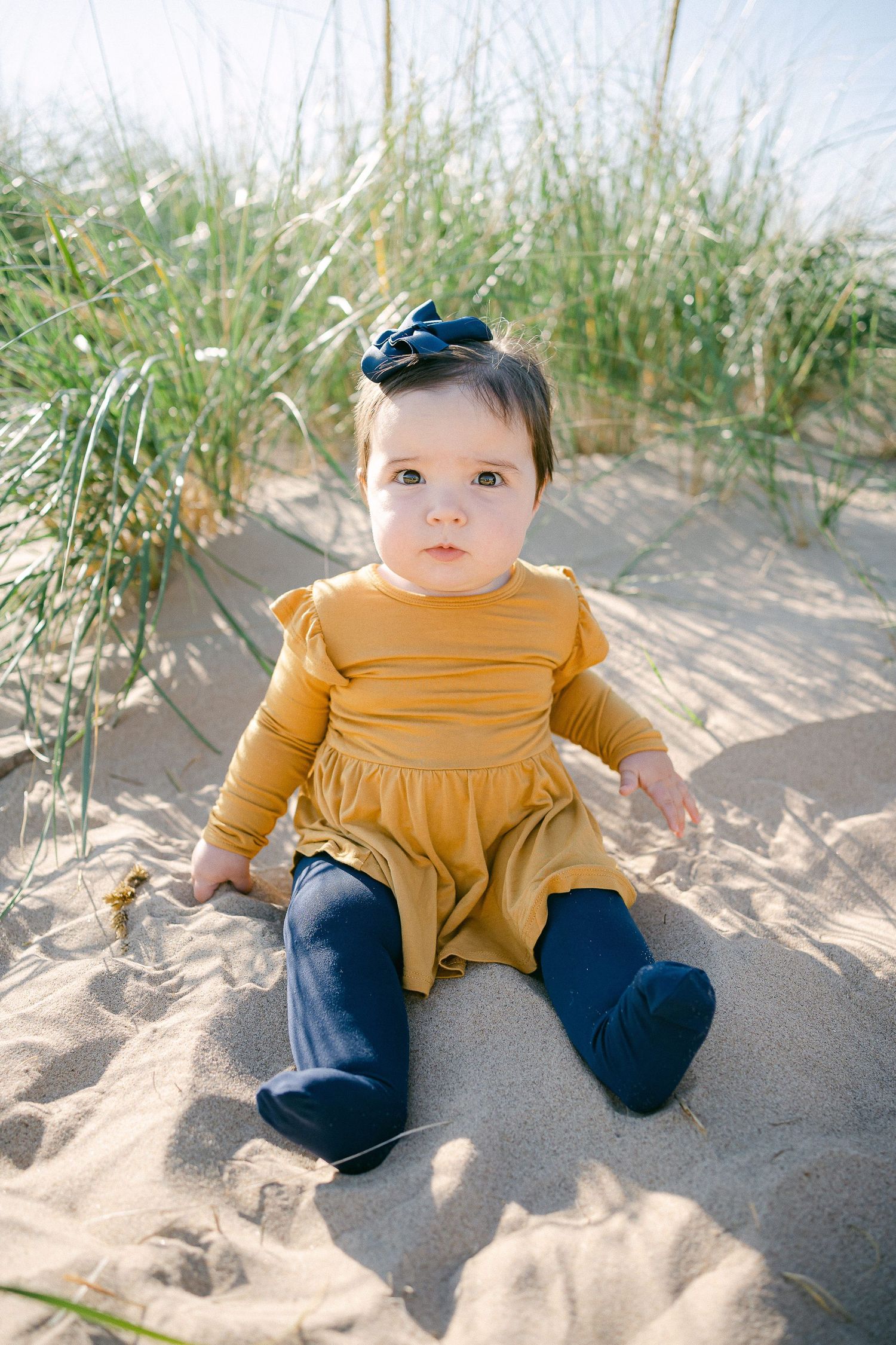 Baby in mustard yellow outfit sits on sandy beach with beach grass in background.