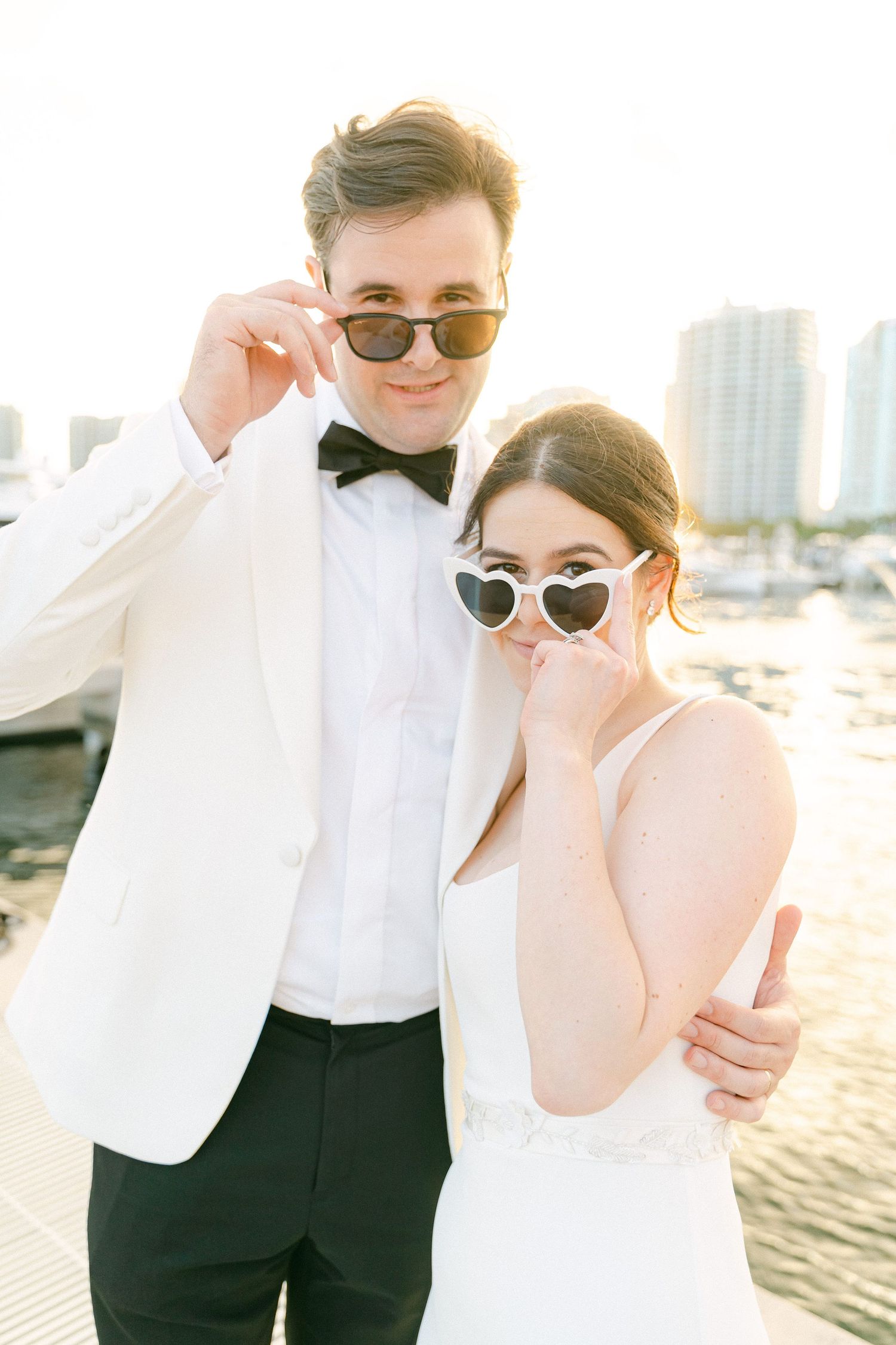 Bride and groom posing on waterfront dock, wearing sunglasses, white tuxedo jacket and heart-shaped frames at golden hour in Miami, FL