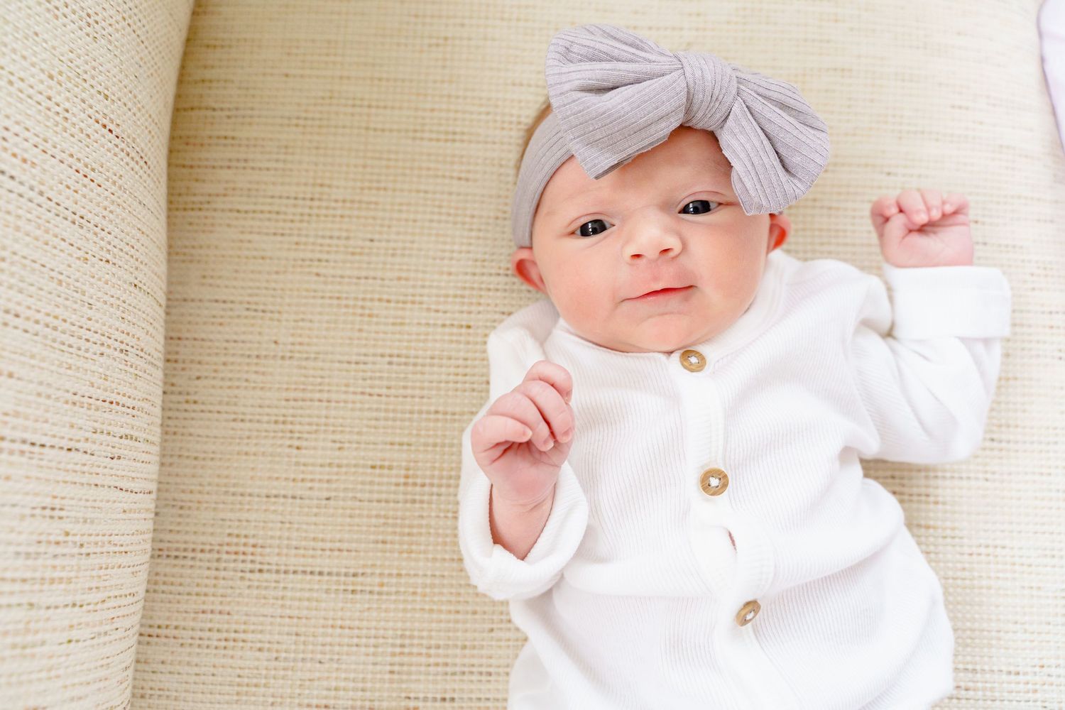 A baby in a white outfit with a bow headband sits and plays on a light-colored textured background.