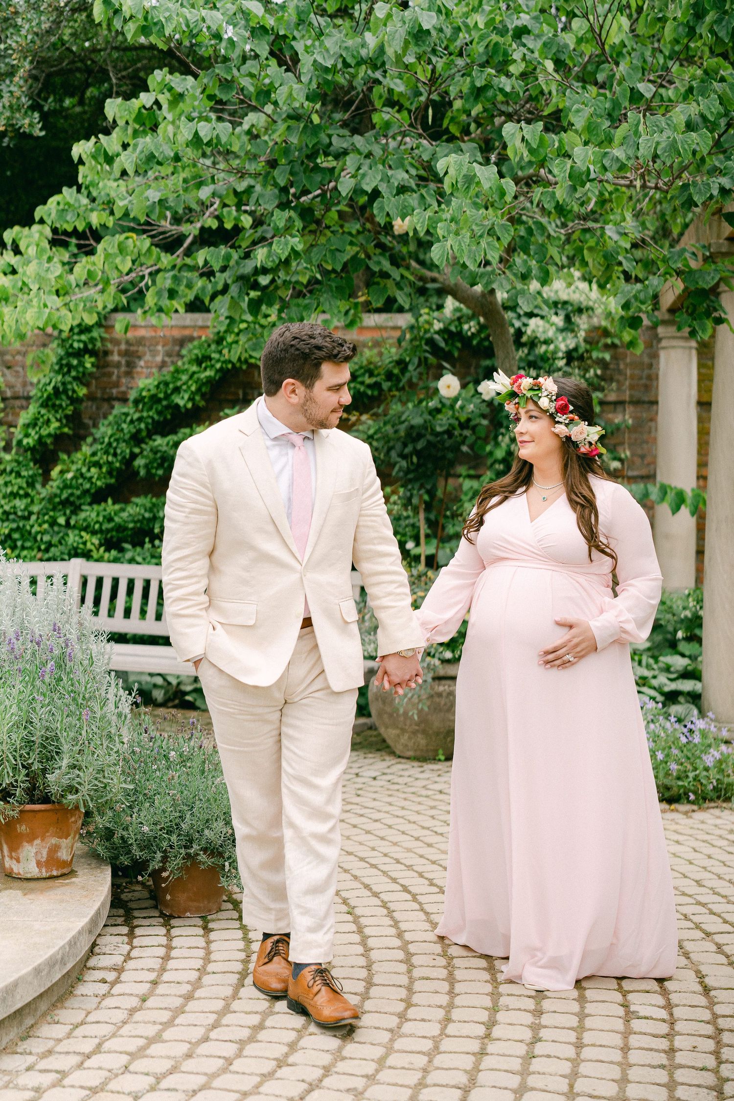 A couple in light colored formal attire poses together on a stone pathway surrounded by lush garden greenery.