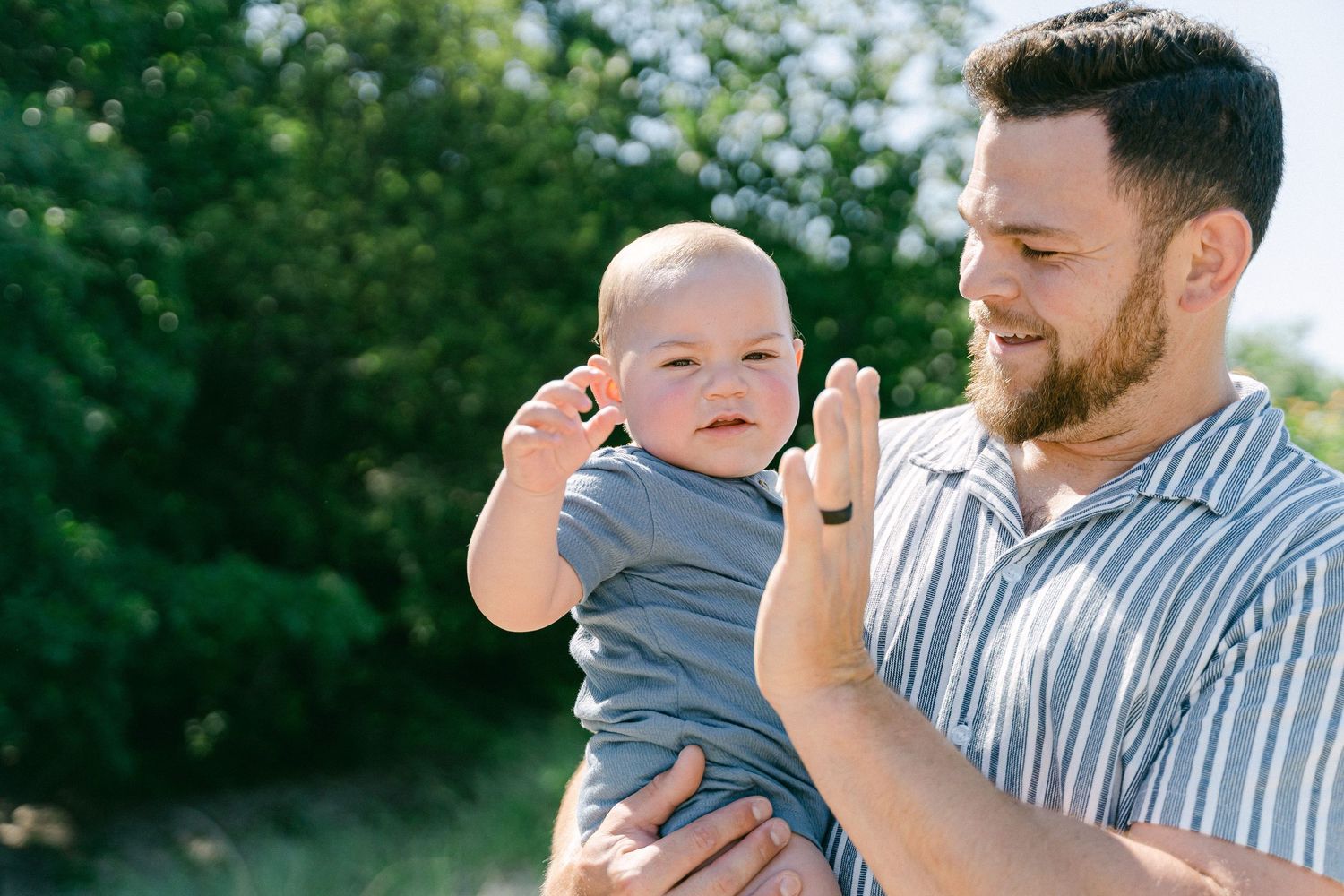 A baby wearing a gray shirt smiles while being held outdoors on a sunny day with green foliage in the background.