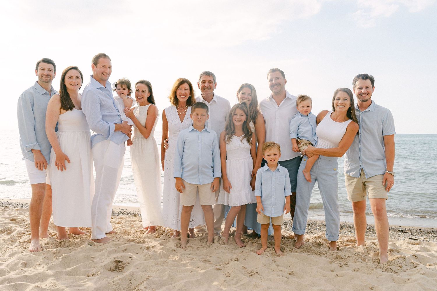 Group of people dressed in light blue and white clothing pose together on a sandy beach with ocean in background.