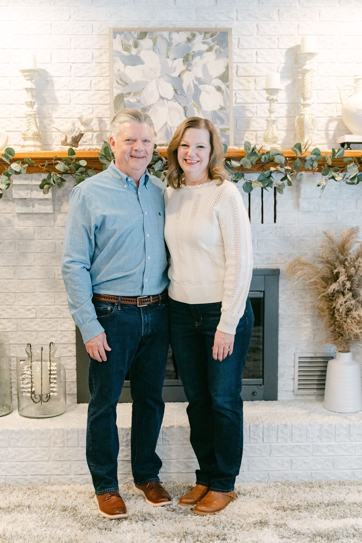 A couple stands together in front of a white brick fireplace decorated with holiday greenery and decor.
